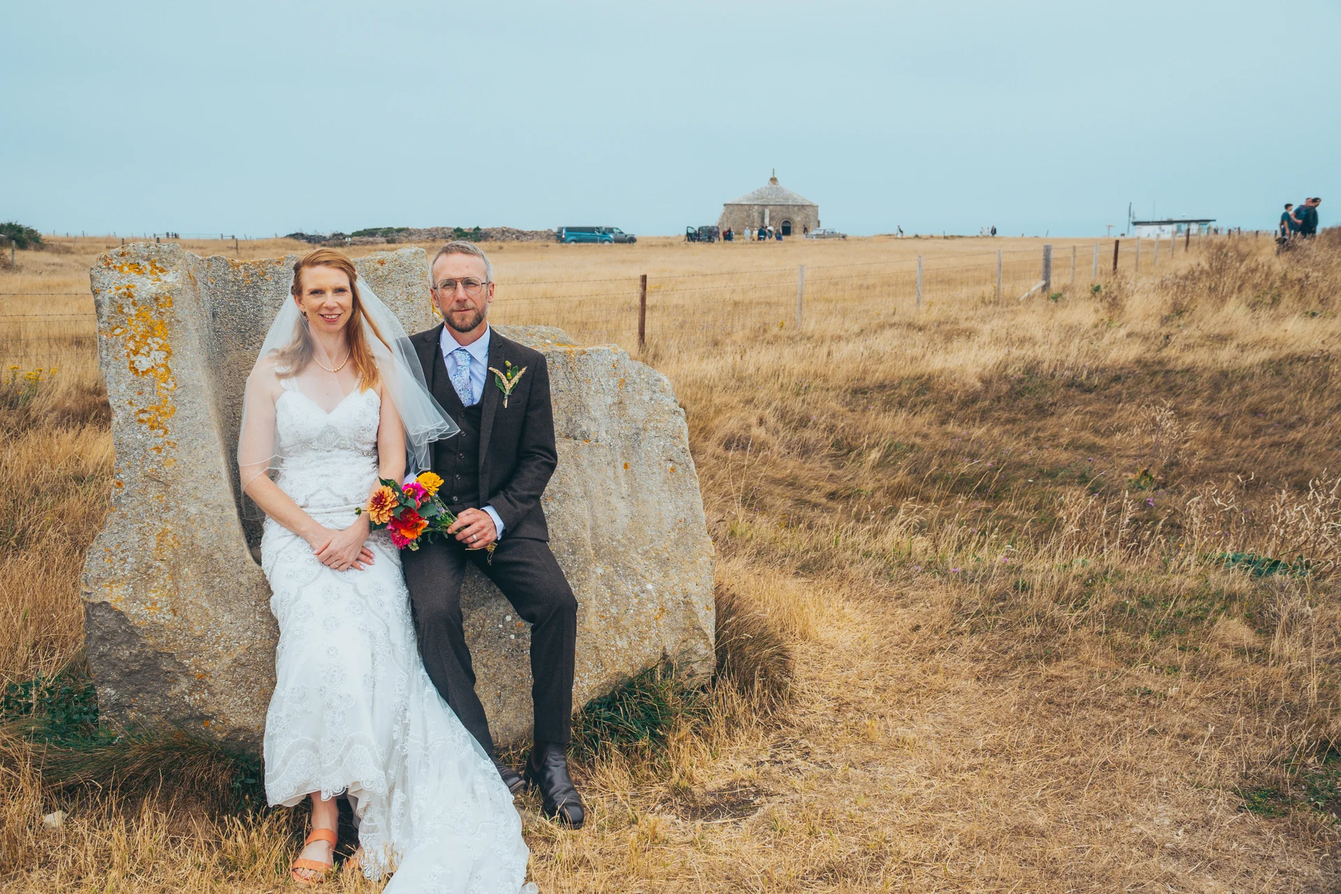 Newlywed bride and groom pose together seated on a large stone monument amidst a vast coastal landscape under an overcast sky.