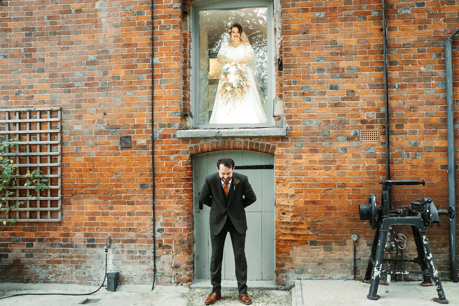 A groom in a green suit stands smiling outside a rustic brick building beneath a window where the bride is visible holding her bouquet.