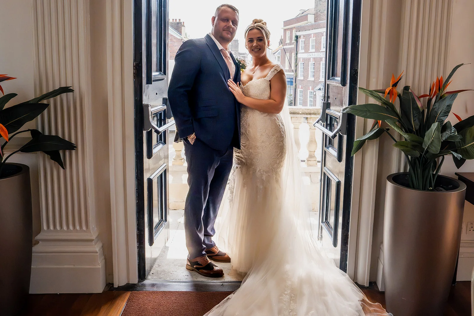 A newlywed couple poses together smiling warmly while standing framed inside an open doorway of a city wedding venue.