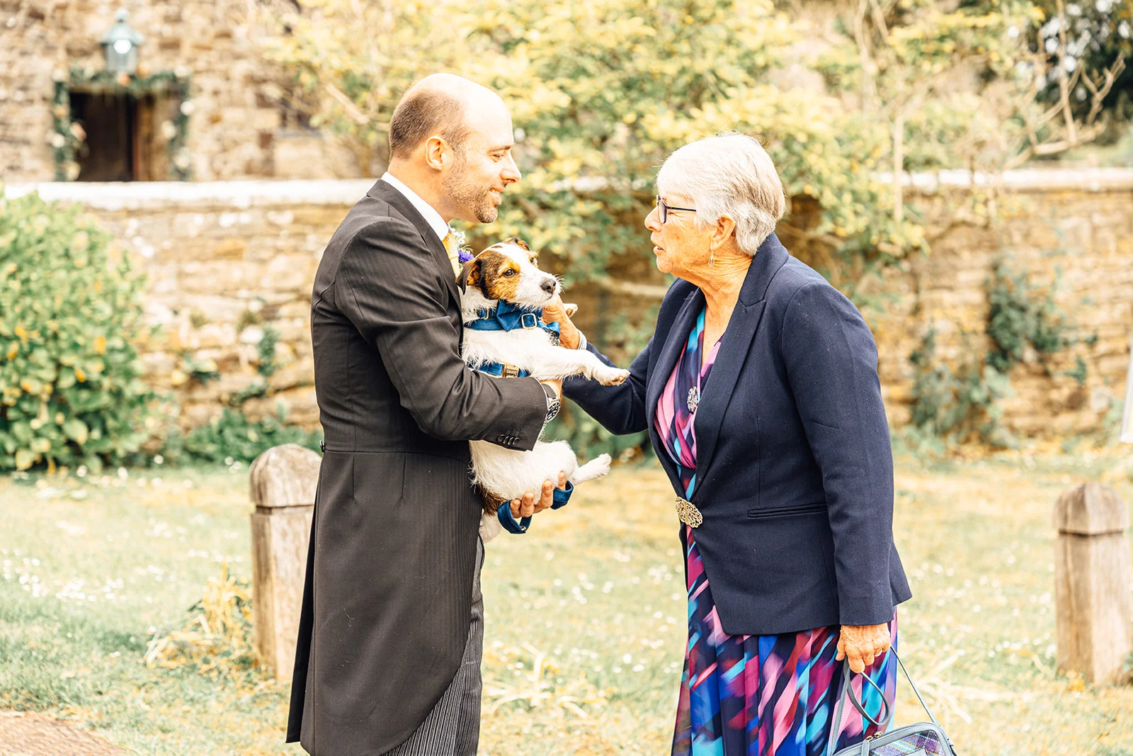 A groom wearing a morning suit holds a small terrier dog wearing a blue bow tie while chatting and laughing with an older guest outdoors near a stone building.