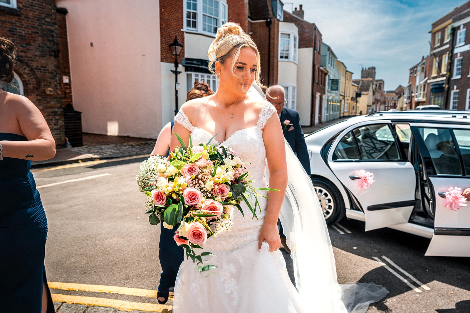 A smiling bride holding a lush floral bouquet walks along a sunlit cobbled street next to a wedding car on her big day.