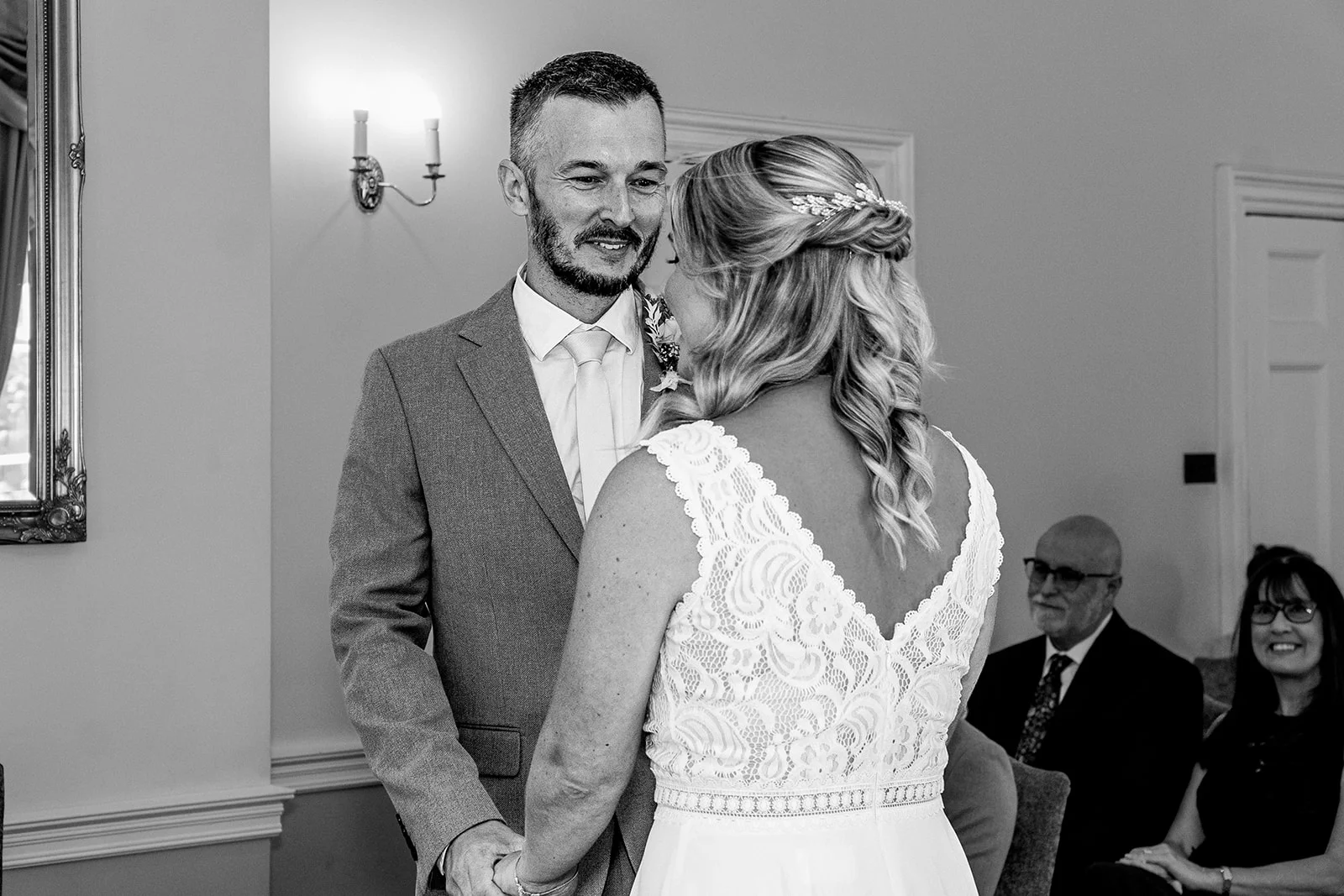 The groom looks emotionally at his bride during the ceremony while holding hands in a classic black and white photograph.