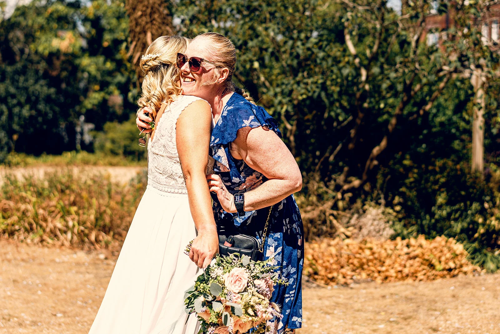 A joyous wedding guest wearing sunglasses and a blue floral dress embraces a laughing bride holding a rustic bouquet outdoors in the sunshine.