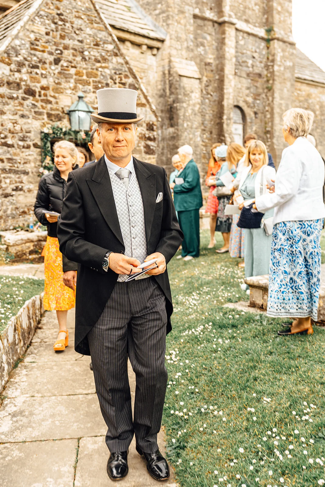 A distinguished guest dressed in a traditional morning suit and top hat stands on a stone path outside a historic church, holding order-of-service booklets while wedding guests mingle in the background.