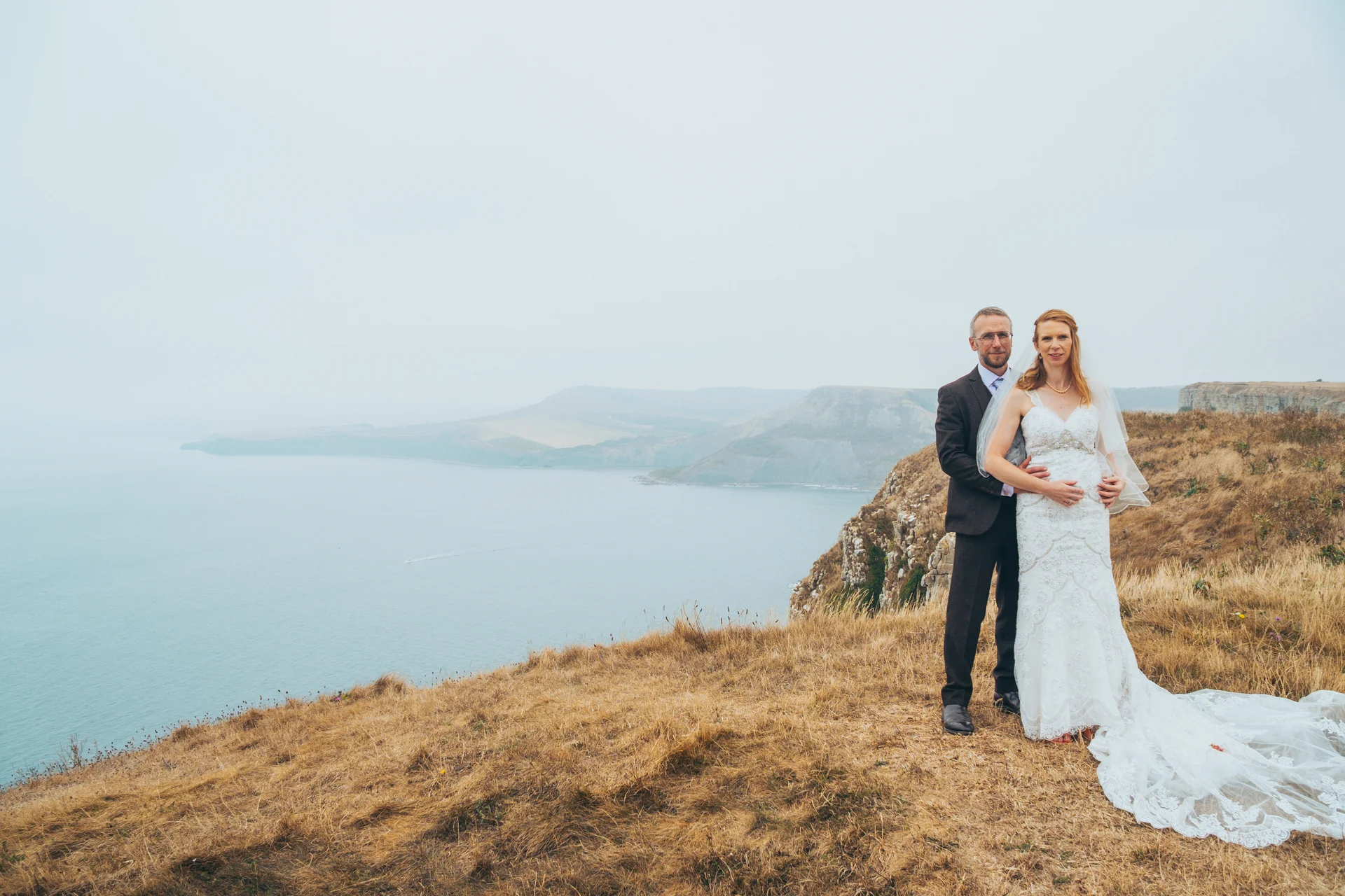 Newlywed couple posing together on a grassy clifftop overlooking the sea on an overcast wedding day.