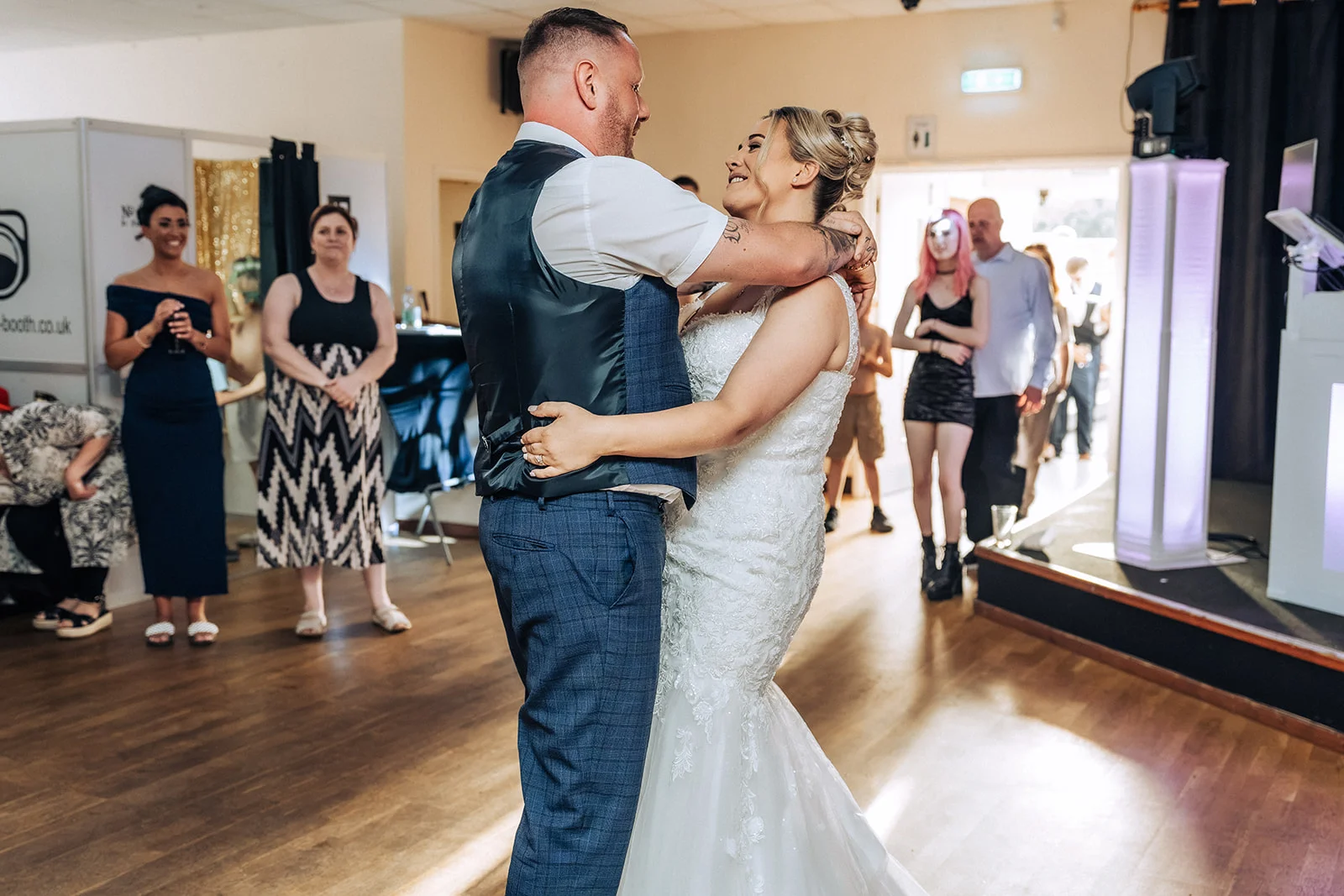The bride and groom share their first dance together on a wooden floor inside a venue while wedding guests watch lovingly from the sidelines.