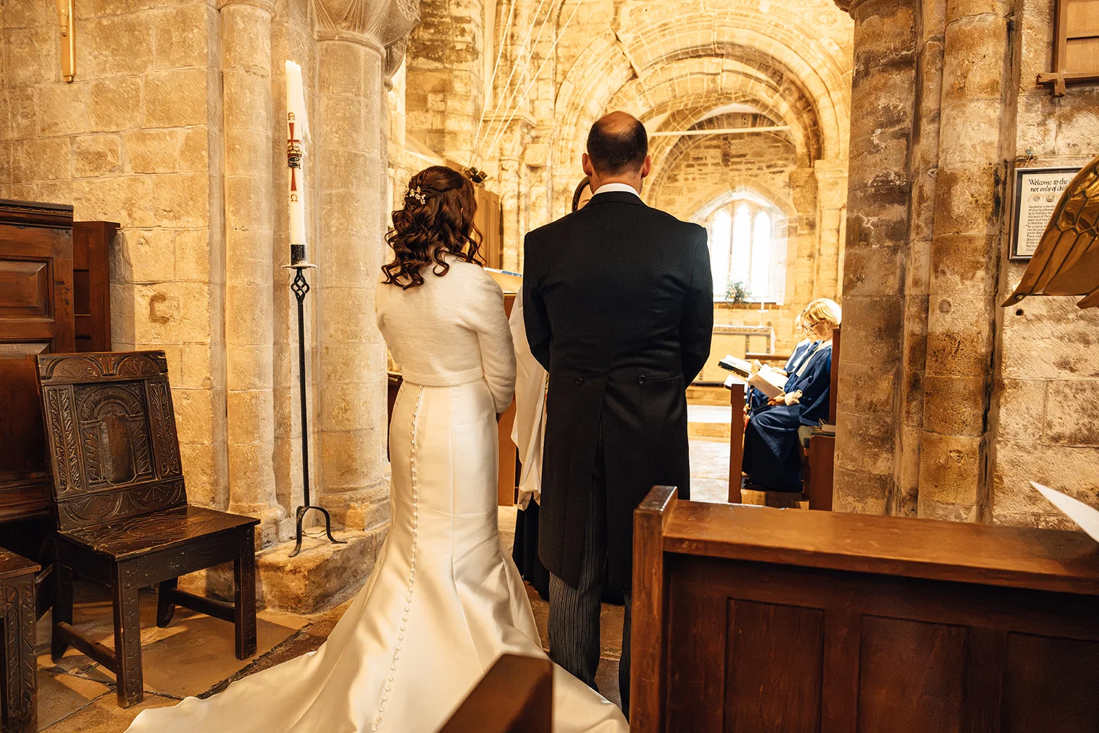 The bride and groom stand together facing the altar during a traditional church wedding ceremony inside a historic stone building.