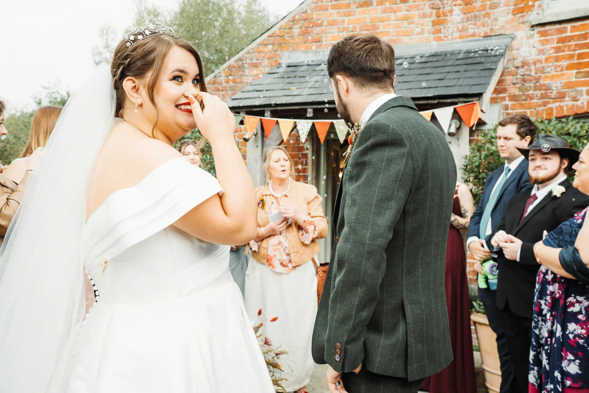 A joyful plus size bride in an off-the-shoulder satin gown and tiara looks back over her shoulder, laughing as she stands outside a rustic brick barn with her groom and wedding guests.