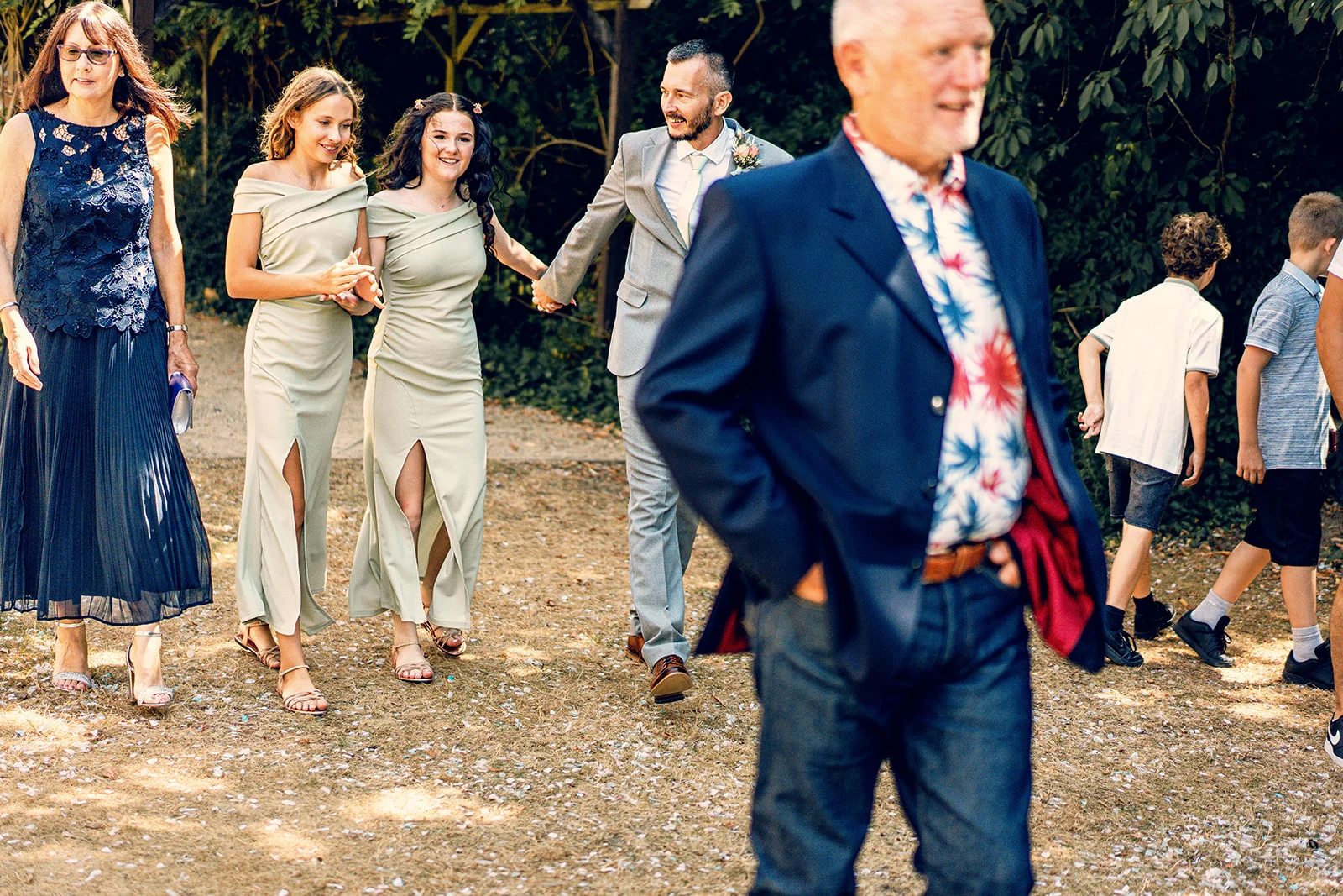 Candid shot of wedding guests and wedding party members walking outdoors on a sunlit path scattered with confetti.