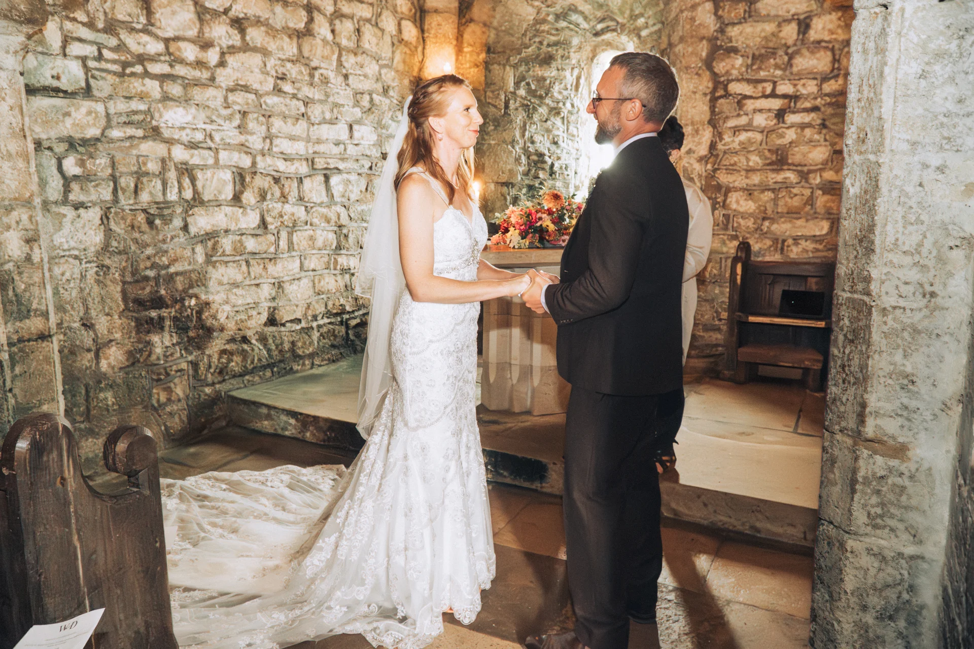 A bride in a beaded fit-and-flare gown with a veil and a groom in a dark suit stand holding hands during an intimate church ceremony against exposed stone walls.