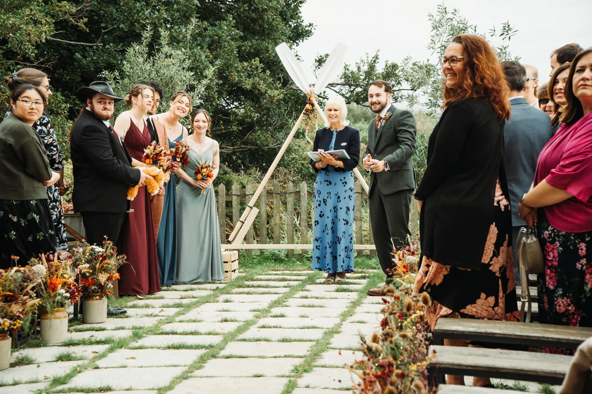 An outdoor wedding ceremony takes place on a paved stone pathway with a celebrant reading vows, a groom in a green suit, and guests watching attentively.