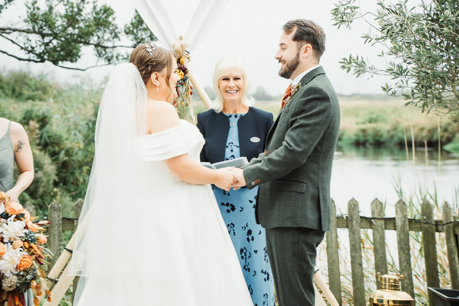 A smiling bride and groom hold hands during an outdoor celebrant-led ceremony by a lake, framed by a tipi structure and rustic wooden fencing.