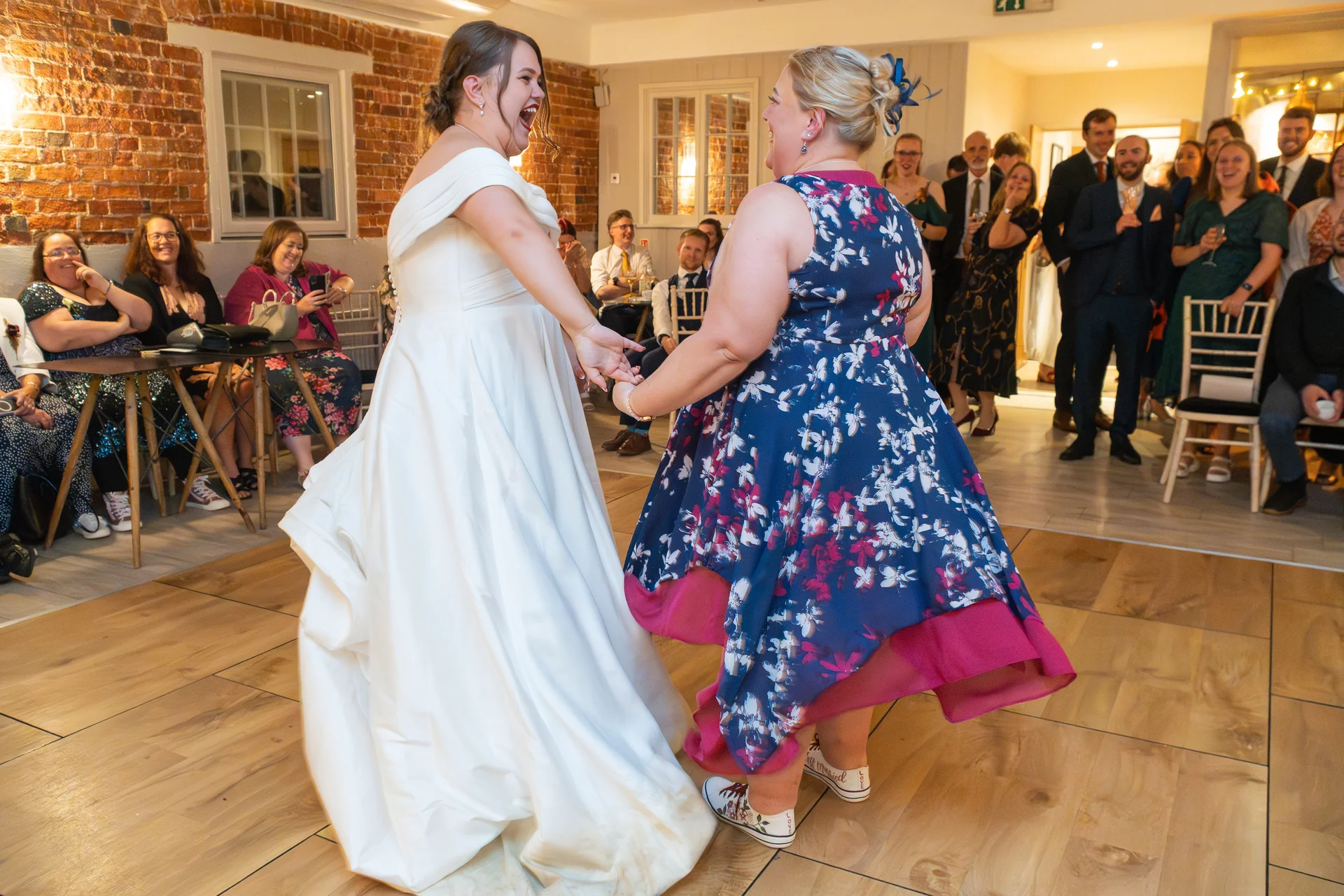 Two brides in wedding attire laugh joyfully while dancing together on a wooden dance floor in an exposed brick venue, surrounded by cheering guests.