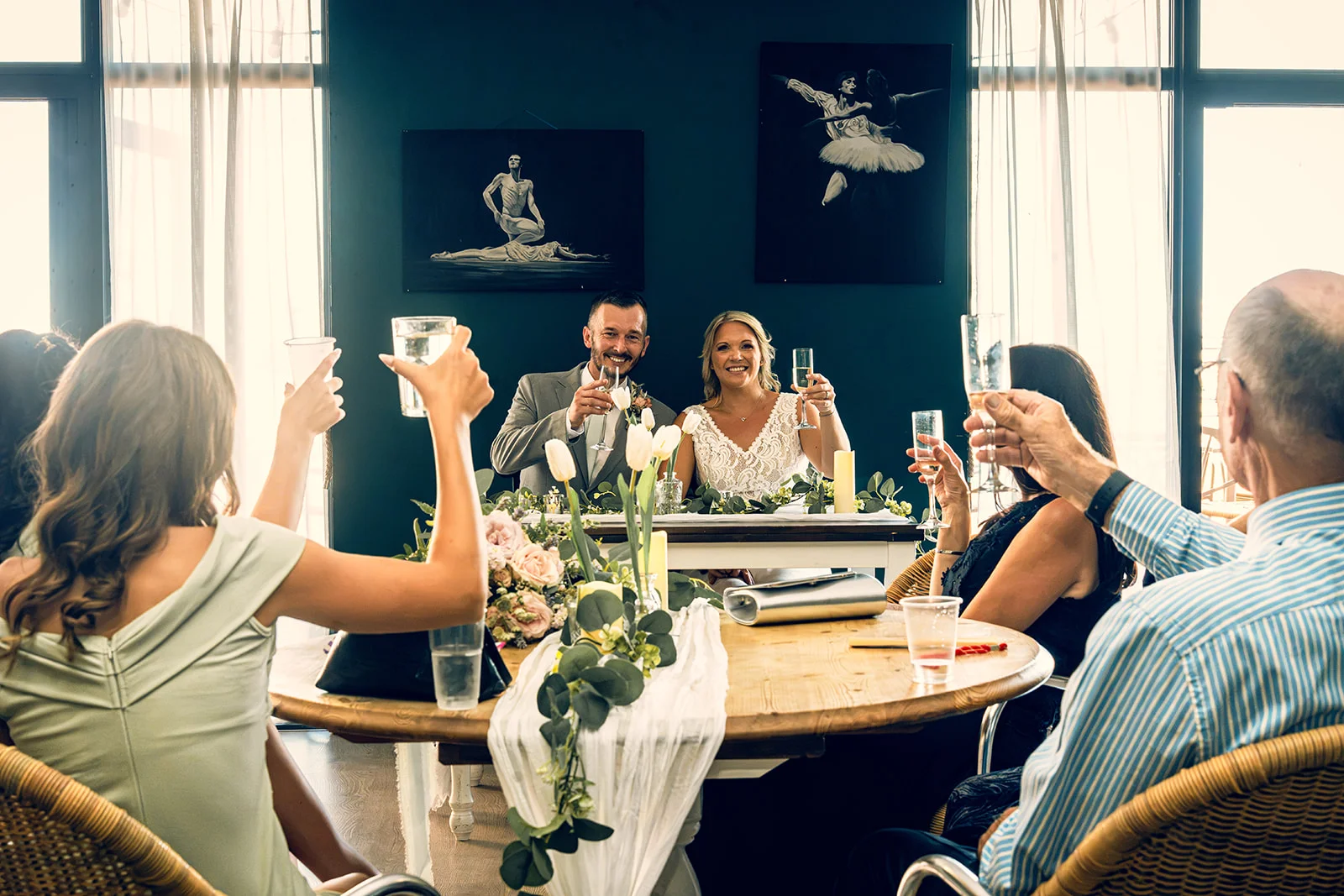 Family and friends raise a toast with champagne flutes at a wedding breakfast, looking towards the bride and groom seated at the top table.