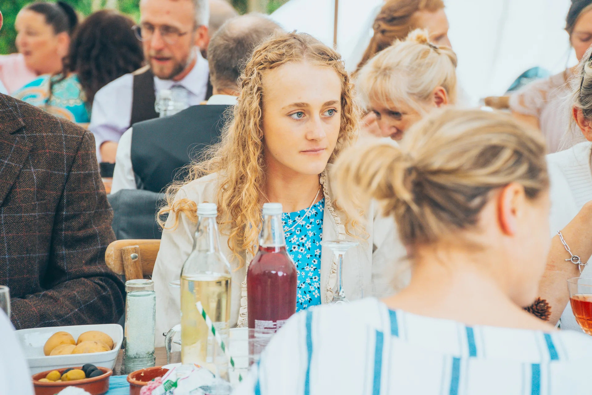 Guests enjoy a wedding breakfast seated around a table with drinks and dishes, with a blonde woman looking thoughtfully towards the side.