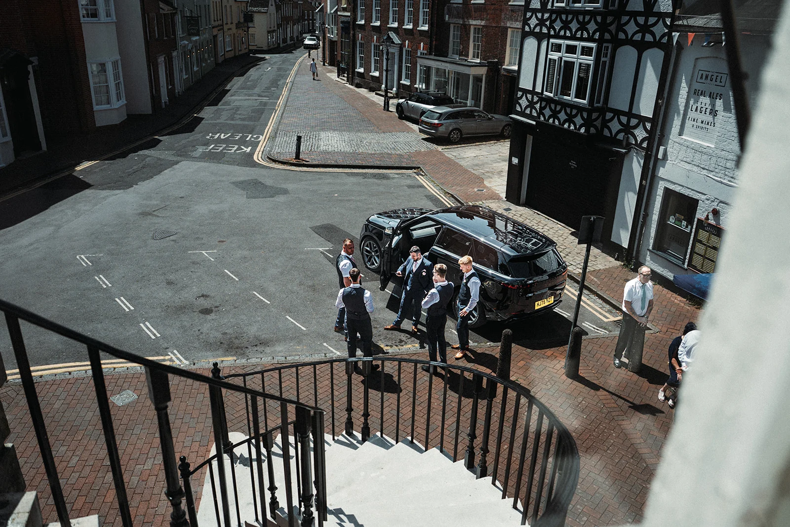An elevated aerial view captures a group of groomsmen gathered around a black luxury car parked on a historic cobbled street outside a traditional English pub.