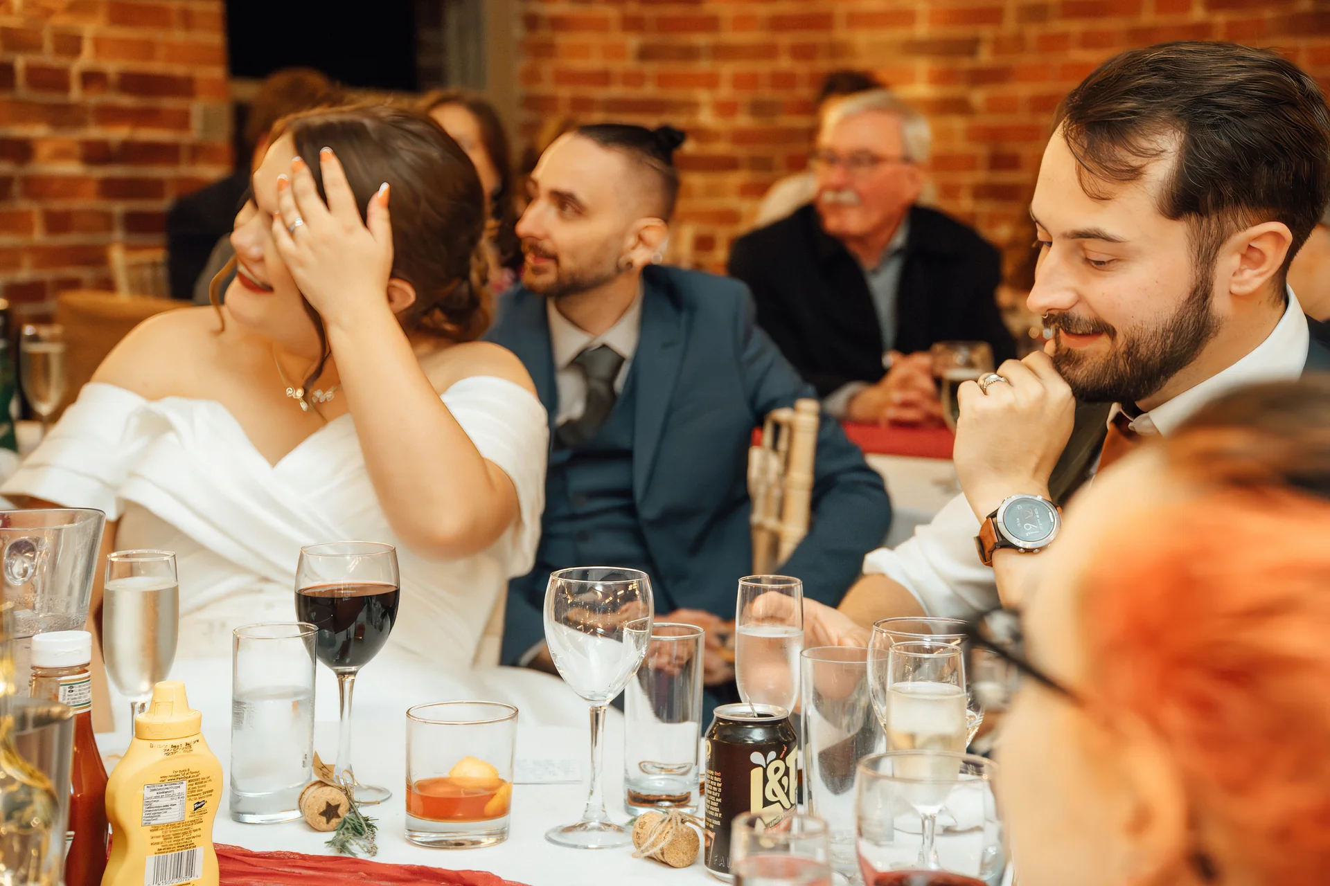Wedding guests laugh and smile during speeches at a reception table set against an exposed brick wall.