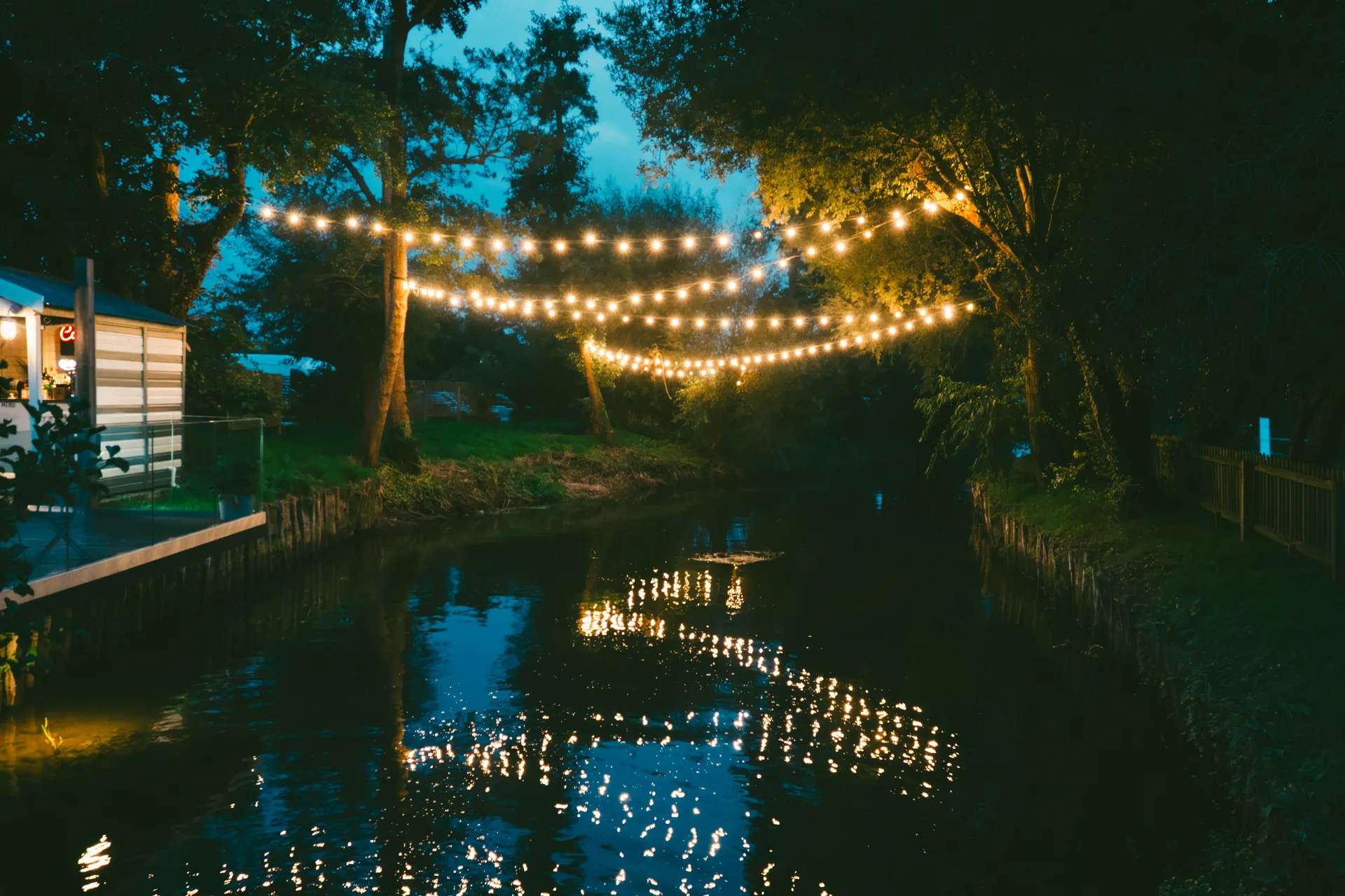A serene riverside wedding venue at dusk, illuminated by glowing festoon lights strung between trees with their reflections shimmering on the water.