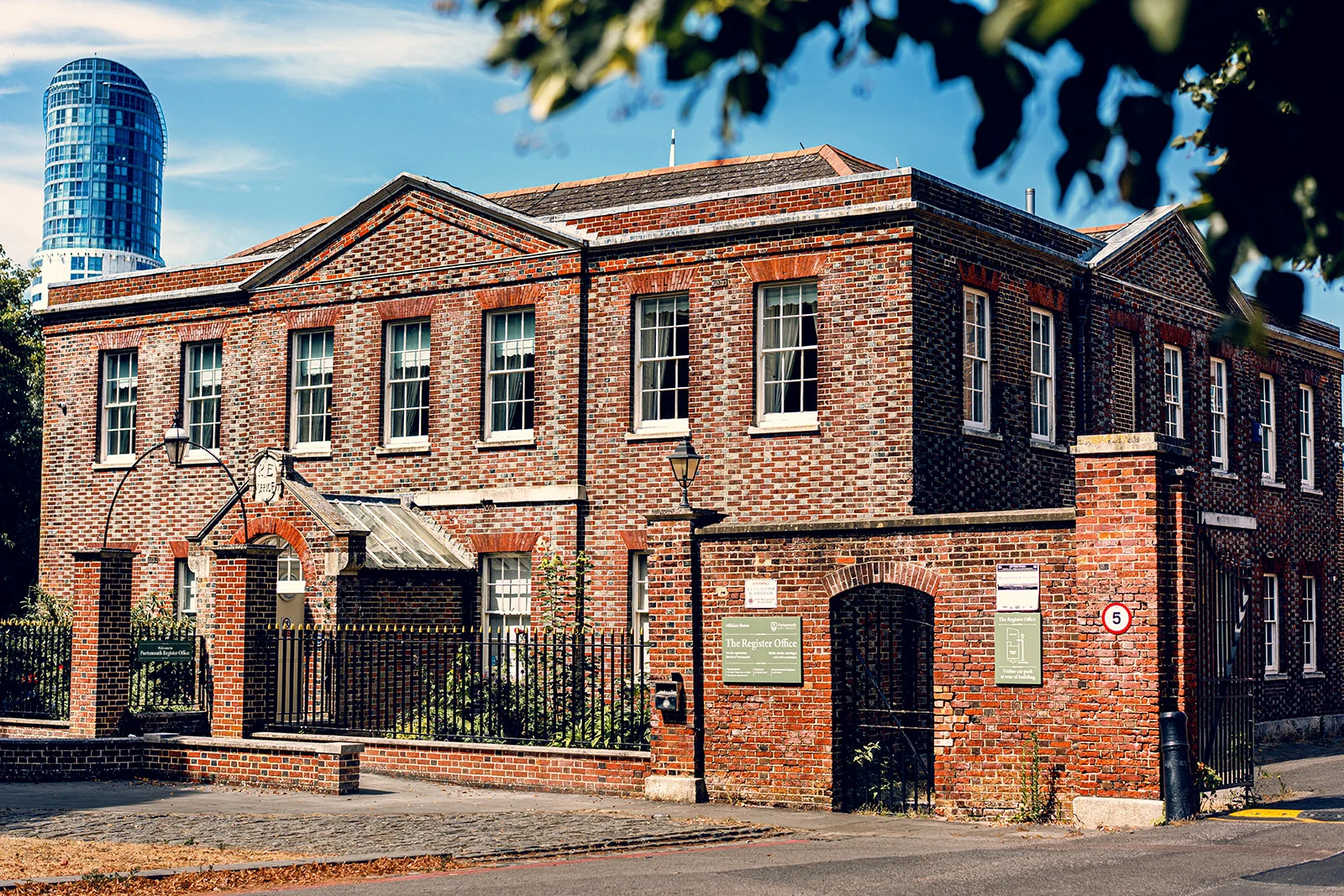 A historic red brick registry office building is illuminated by bright sunlight under a clear blue sky, featuring a modern glass skyscraper in the distant background.
