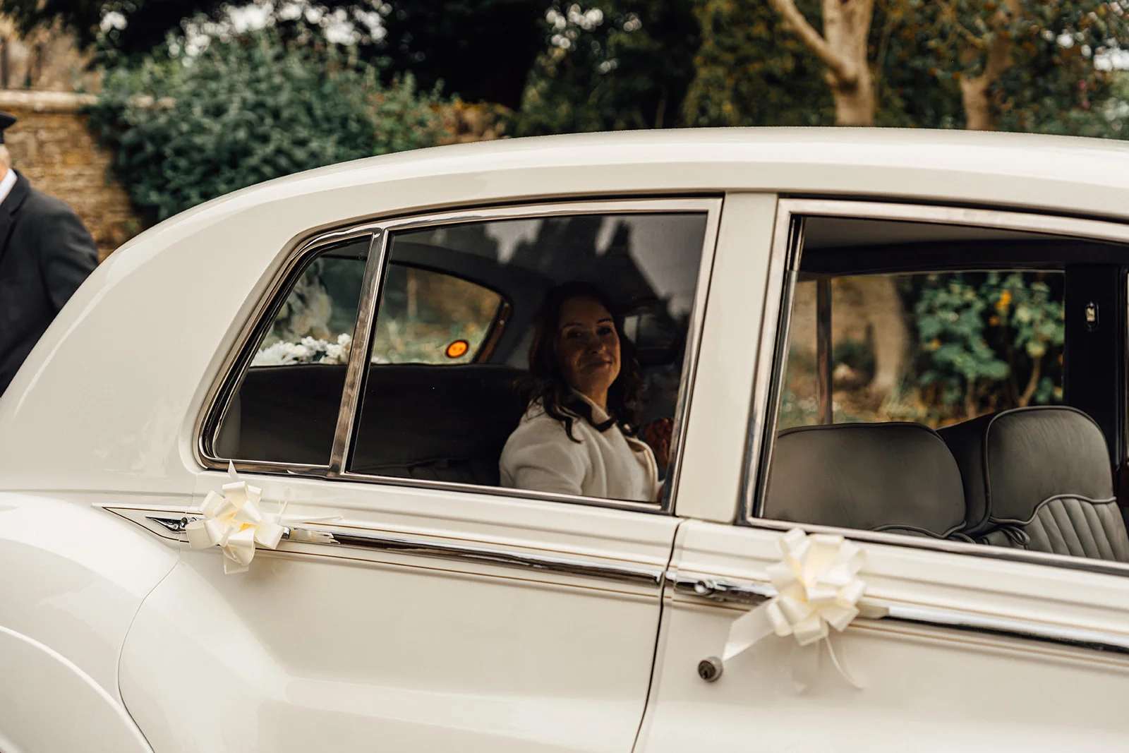 A smiling bride looks out from the back seat of a classic white wedding car decorated with white ribbons on the door handles.