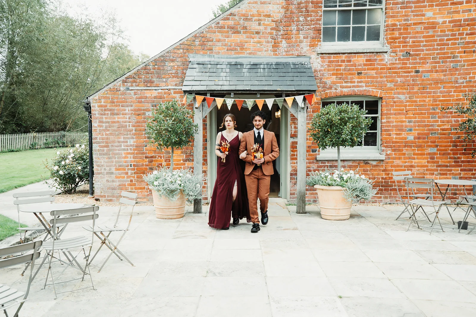 A bride in a striking burgundy dress and a groom in a tailored brown suit walk arm-in-arm out of a rustic red brick barn adorned with colourful bunting.