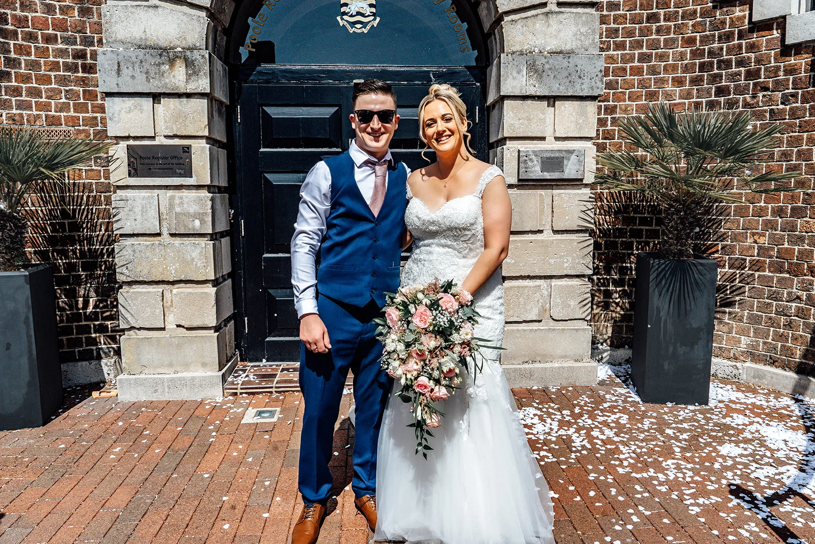 A newlywed couple poses outside a historic brick building with stone archways, scattered with white confetti petals on the brick paving.