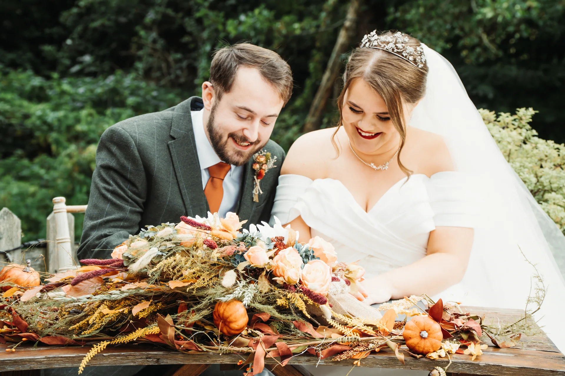 A smiling bride and groom sit together outdoors signing their marriage register, framed by an abundant autumn-themed floral arrangement with mini pumpkins and foliage.