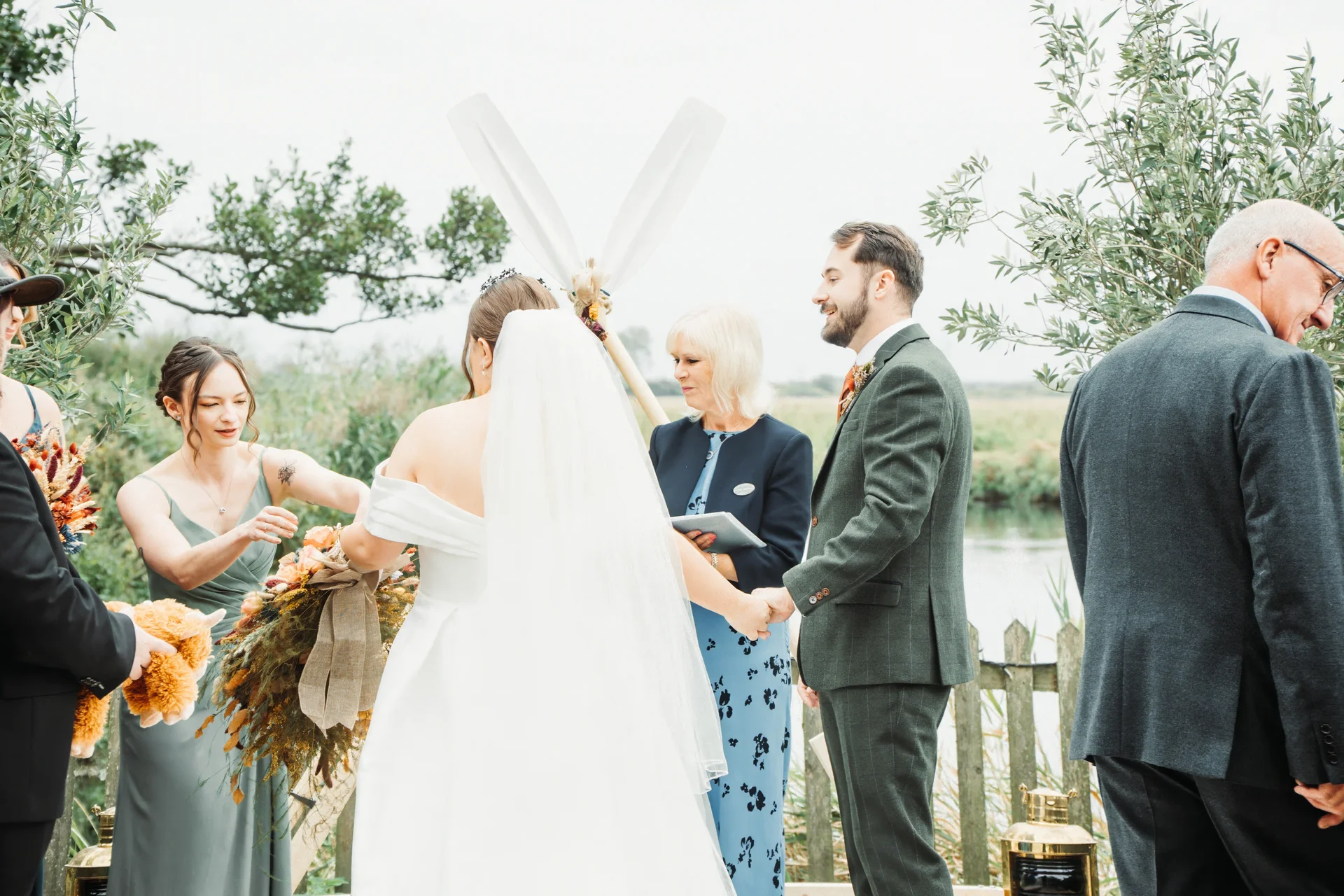 A bride and groom participate in an outdoor, celebrant-led ceremony by a river, with the groom wearing a green suit and the bride in a classic white off-the-shoulder gown and veil.