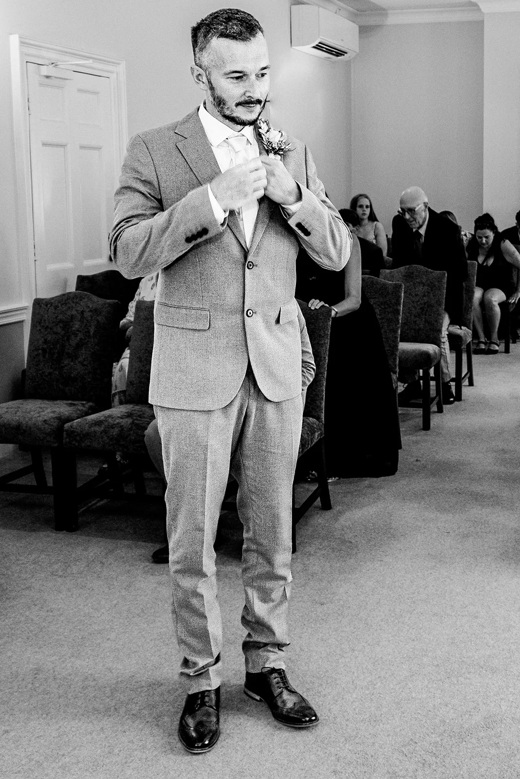 A groom in a light grey suit stands adjusting his lapel at the front of a registry office ceremony room while seated guests look on.
