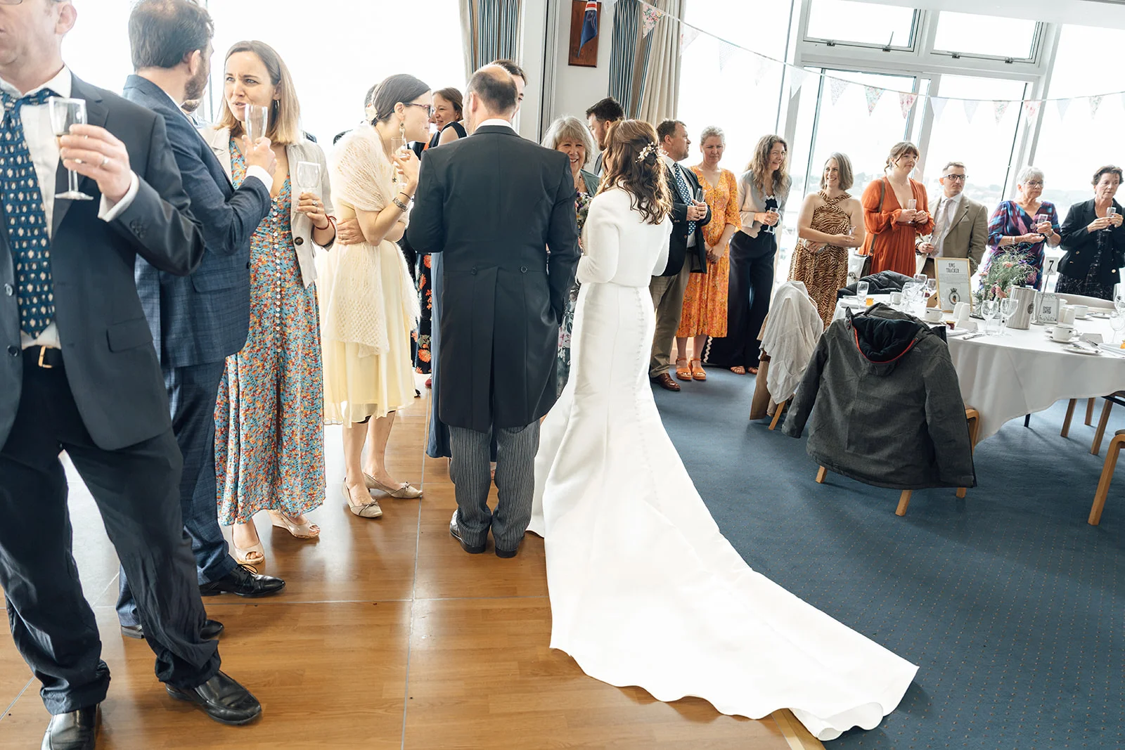 Guests mingle and chat during a bright drinks reception while the bride in a long sleeved white gown and groom in a morning suit stand together.