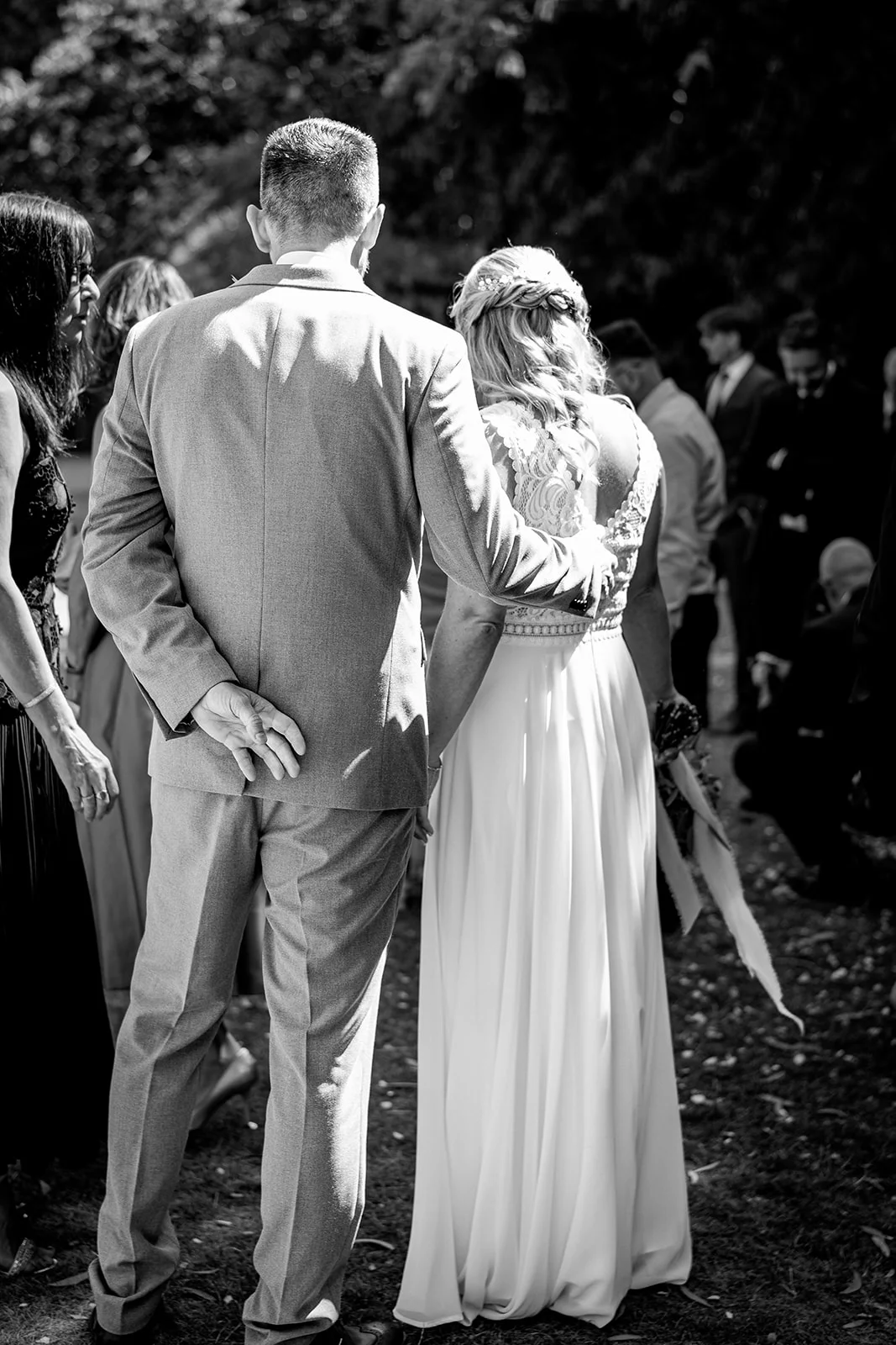 A newlywed couple stands together outdoors from behind during their wedding celebration, with the groom in a grey suit holding the bride's hand.