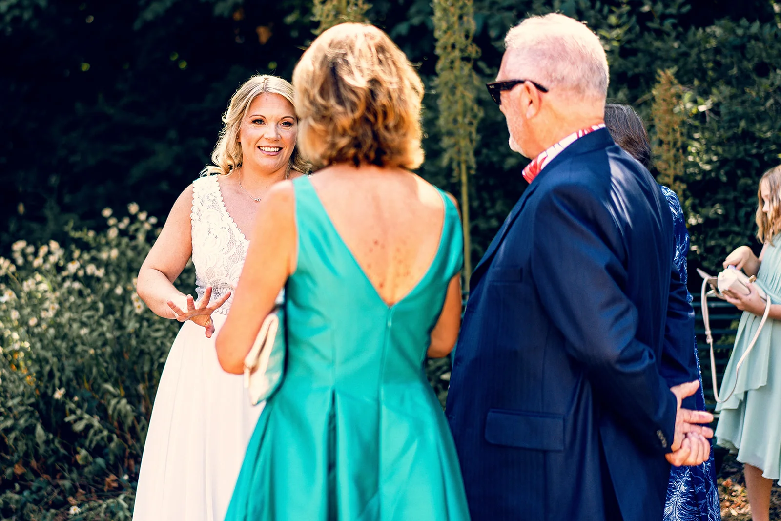 A smiling bride in a lace-detailed wedding dress chats with guests outdoors in a sunlit garden setting during the drinks reception.