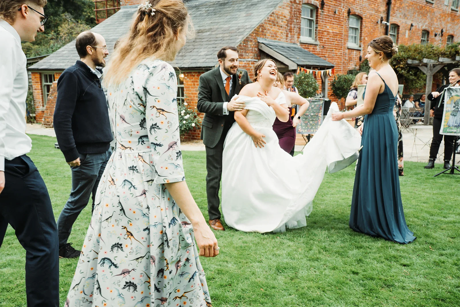 A joyous bride and groom laugh heartily outdoors on a lawn surrounded by guests, with a brick building in the background.
