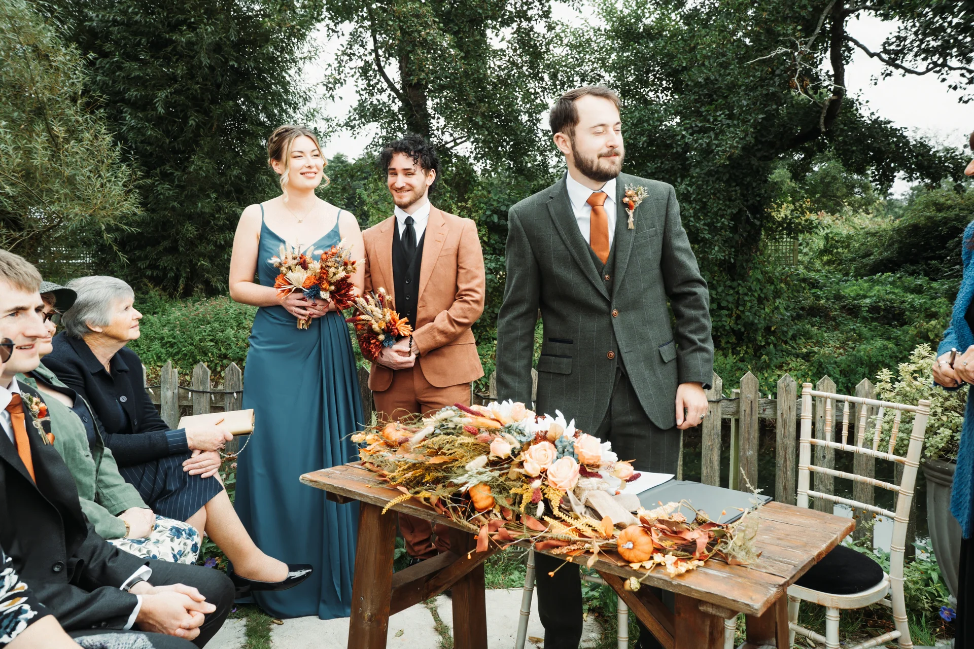 An outdoor wedding ceremony features a groom in a green tweed suit standing beside a rustic wooden signing table adorned with autumn foliage and pumpkins, while guests and wedding party members look on.