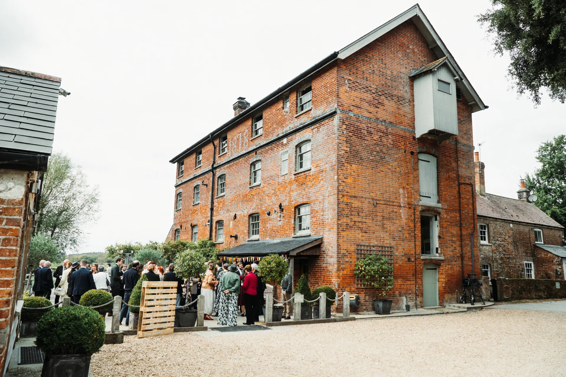 Wedding guests gather outside a large industrial red brick warehouse venue during the daytime drinks reception.