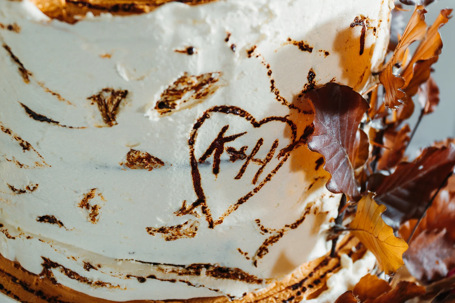 Close-up of a white textured buttercream wedding cake featuring a carved heart with initials and decorated with autumn leaves.