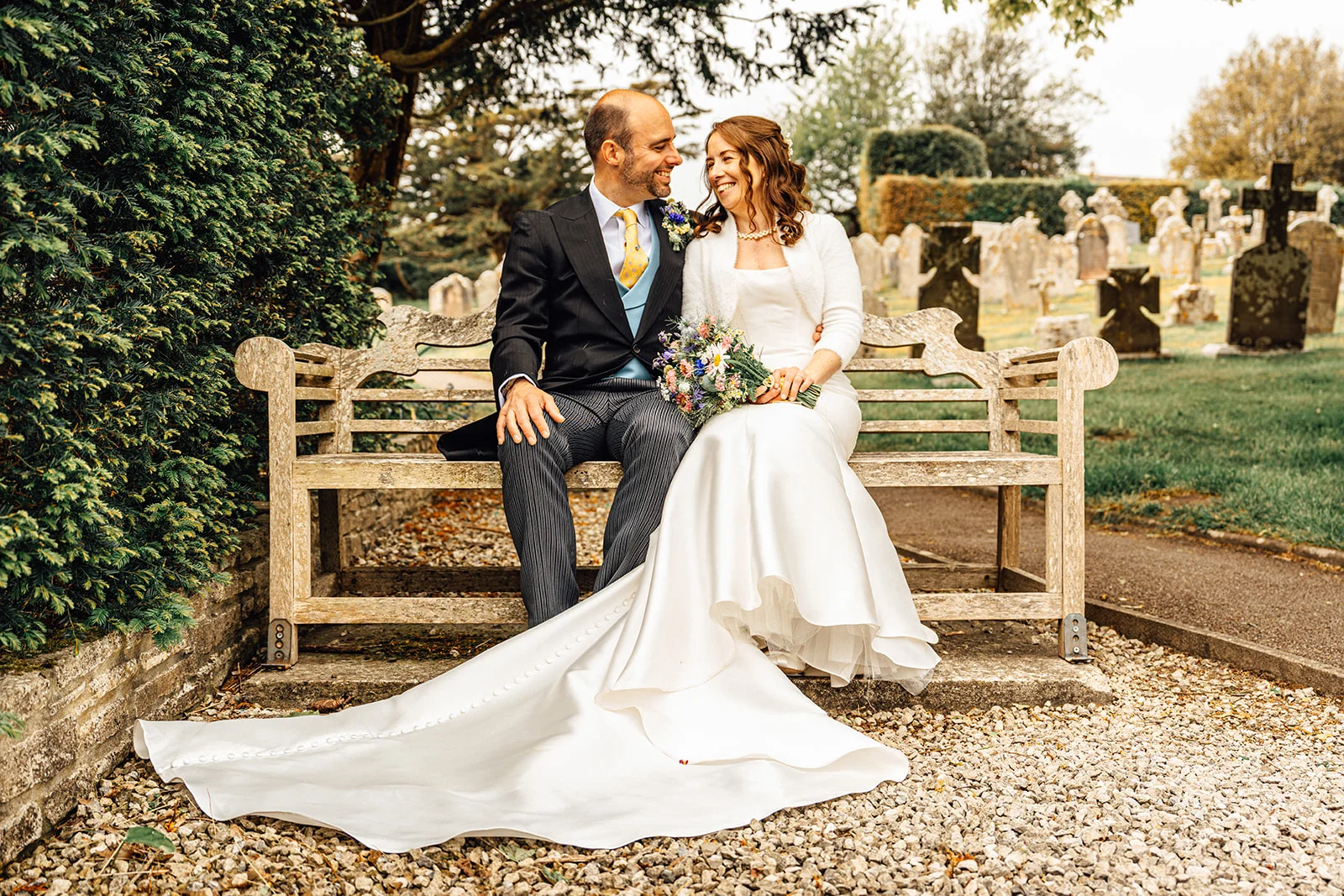 A newlywed couple smiling at each other while seated on a wooden bench outside a historic churchyard.