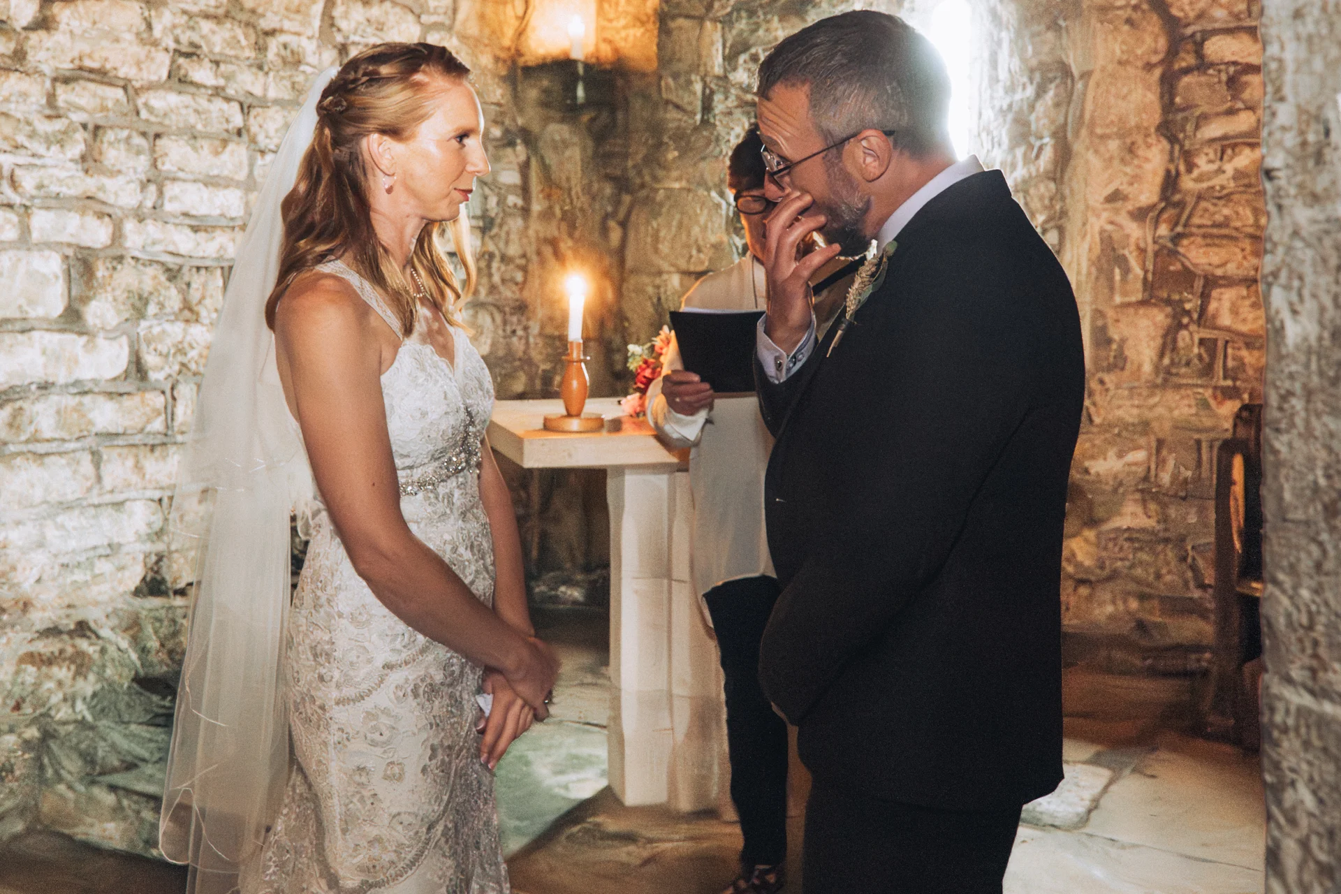 An emotional groom wipes away tears while standing opposite his bride during an intimate candlelit ceremony inside a historic stone building.