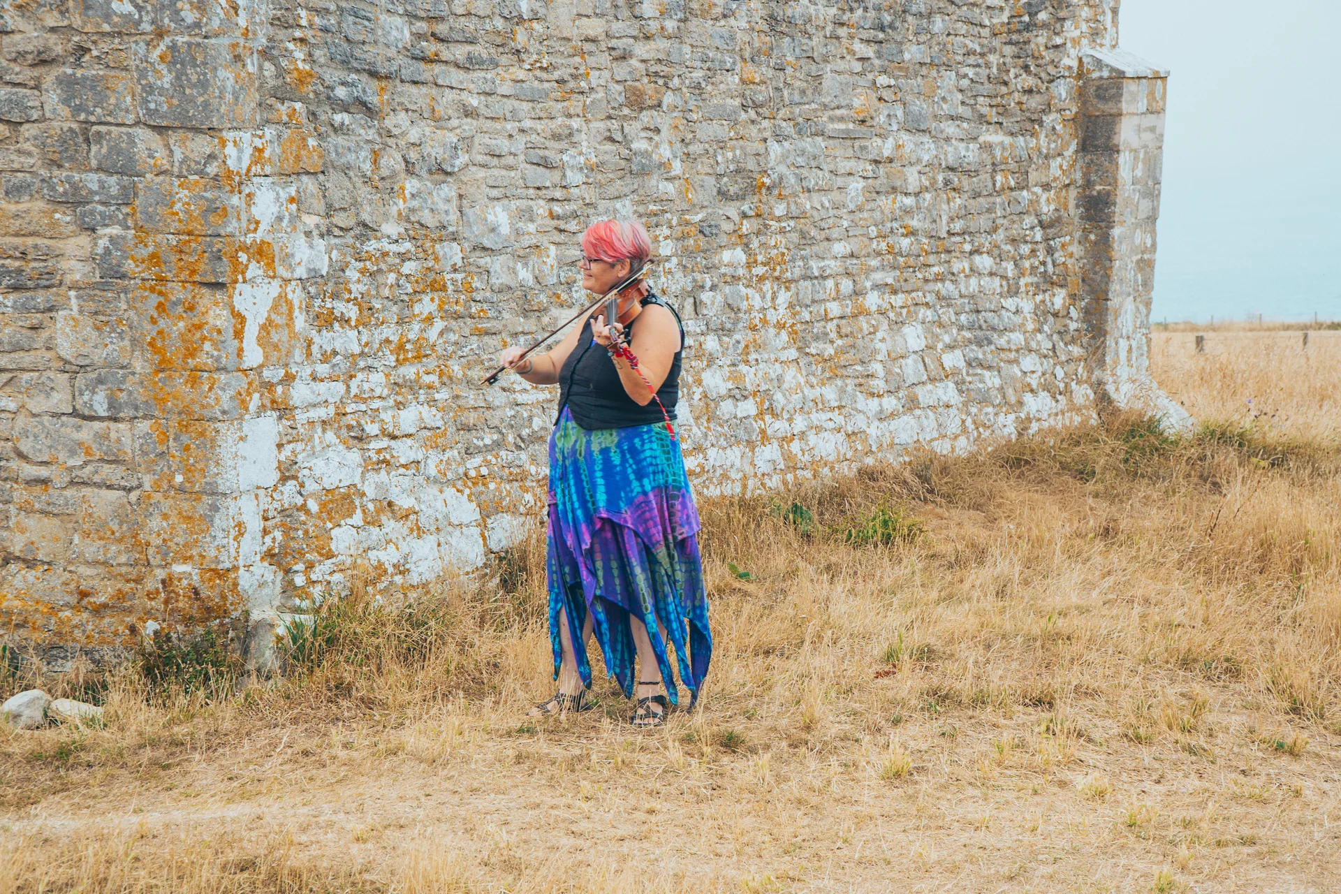 A musician with vibrant pink hair plays the violin outside against a weathered stone wall during an outdoor wedding.