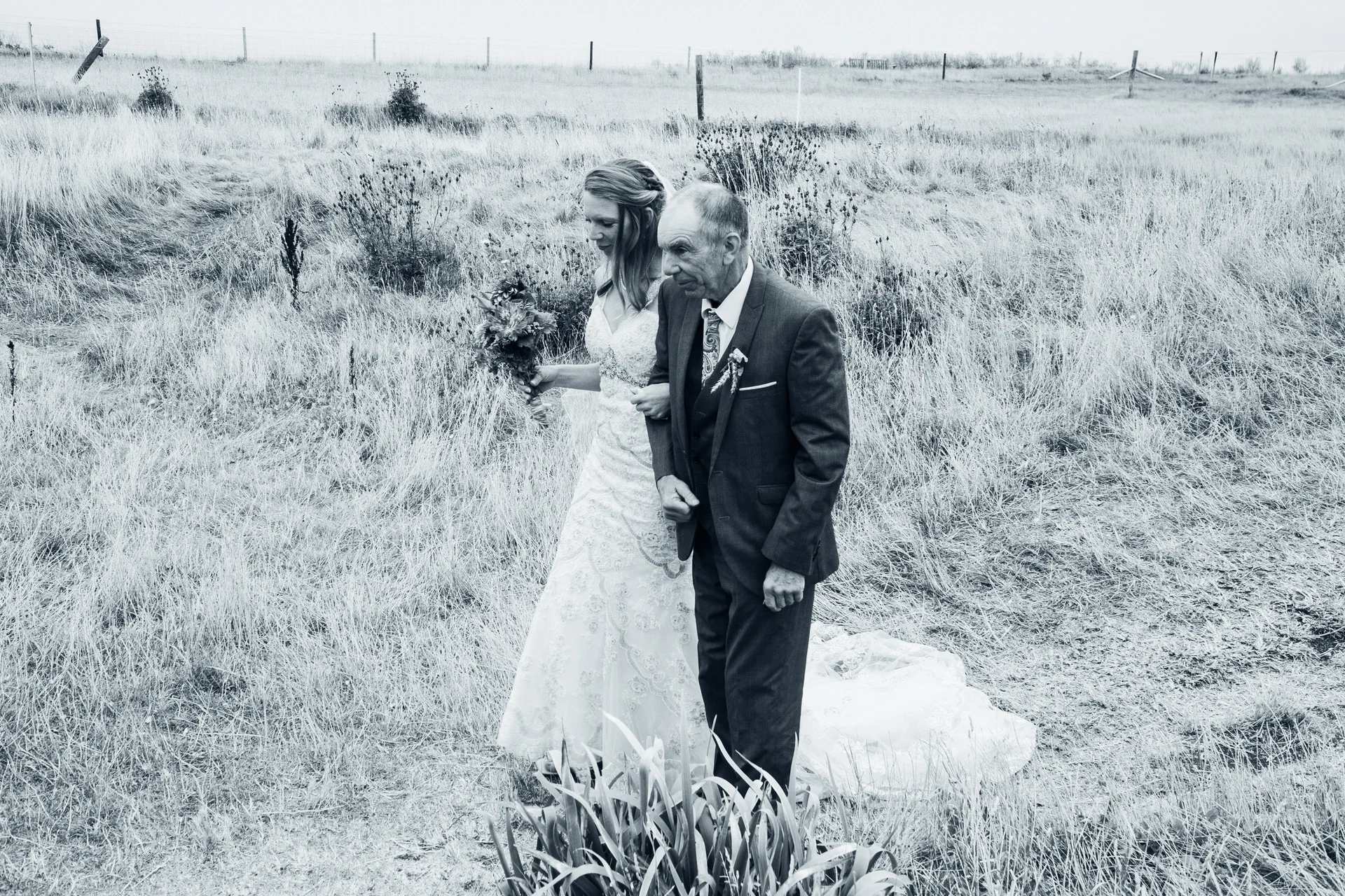 A bride in a patterned lace gown holds a bouquet while walking arm-in-arm with her father through a grassy field in a black and white photograph.