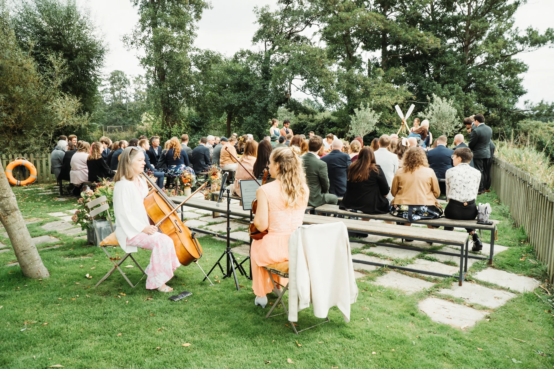 A cellist and violinist perform outdoors on the lawn at an open-air wedding ceremony as guests sit on wooden benches surrounded by lush greenery.
