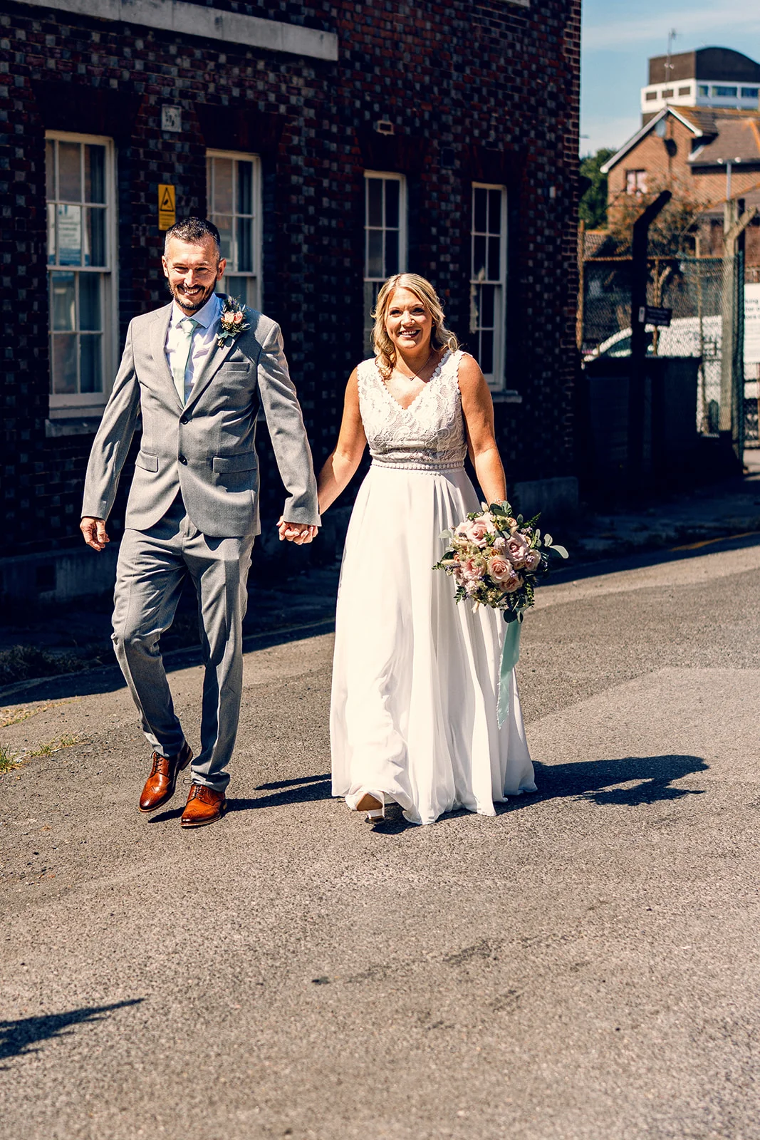 A smiling bride and groom walk hand in hand down a sunlit city street past an exposed brick building on their wedding day.