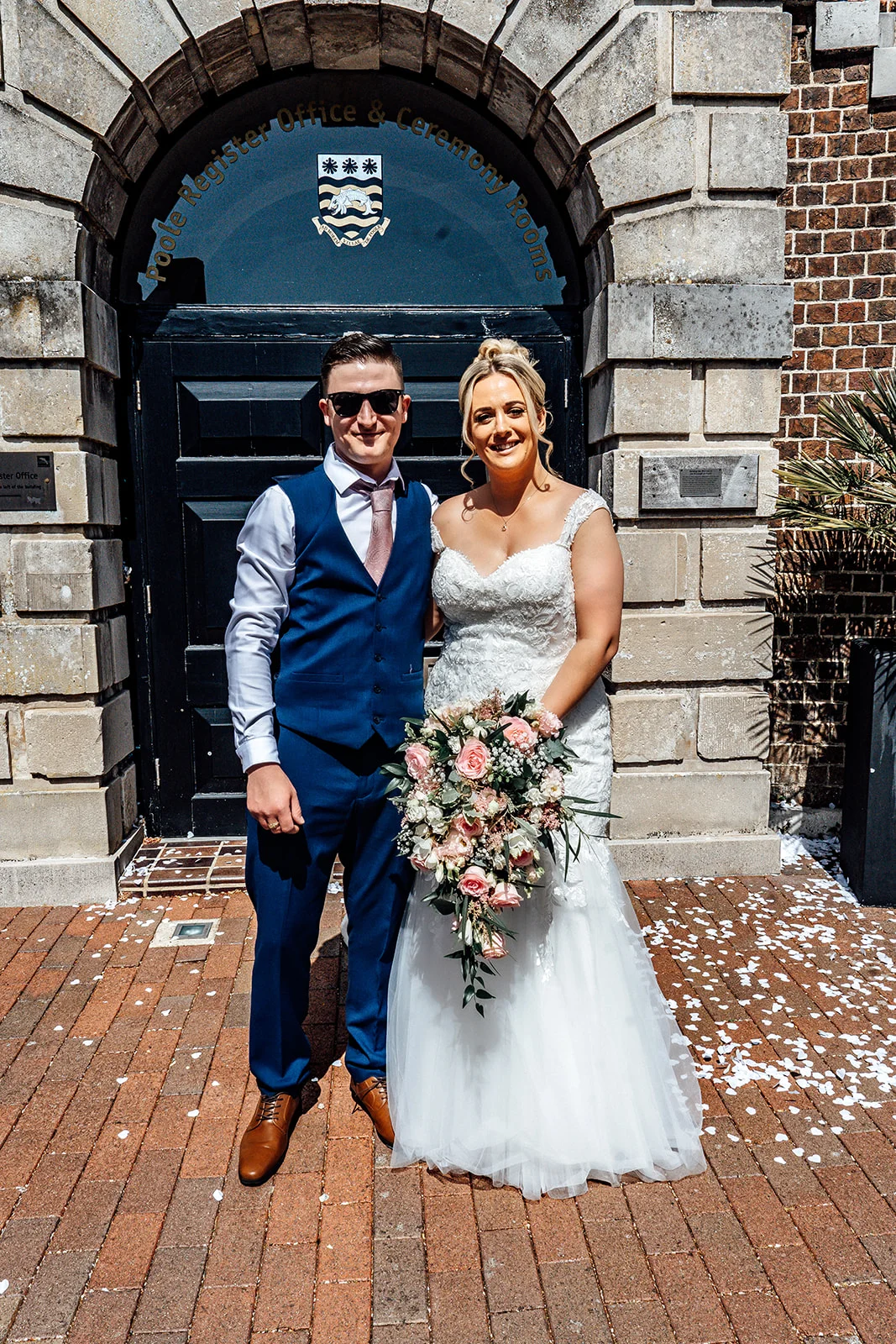 Newlywed bride and groom stand smiling outside a registry office entrance with a stone archway, holding a large cascading floral bouquet amidst scattered white confetti.
