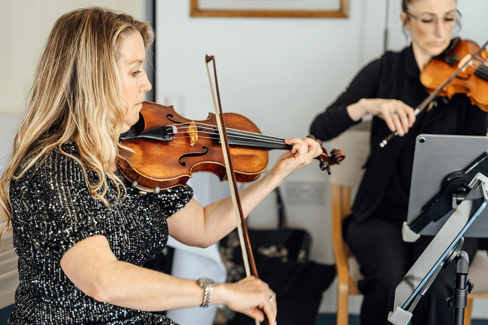 Two female musicians focused on playing their violins during a wedding reception, with the foreground violinist wearing a sparkling black dress.