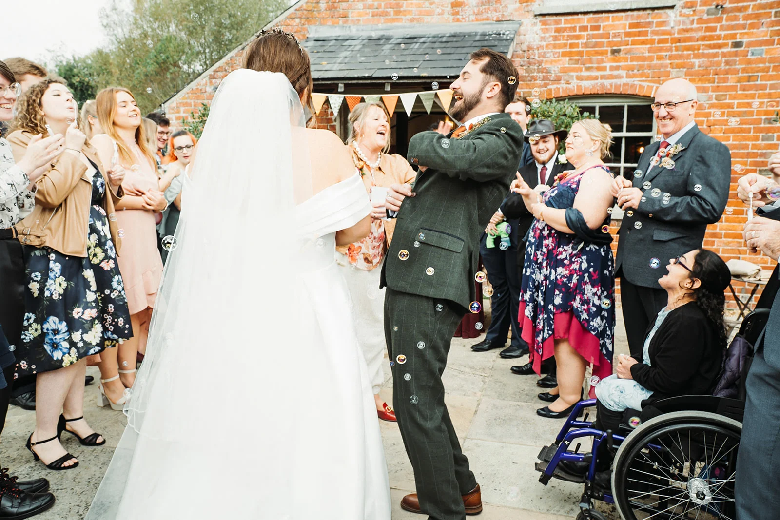 A newlywed couple laugh ecoperatively as guests blow bubbles during their exit outside a rustic brick barn venue.