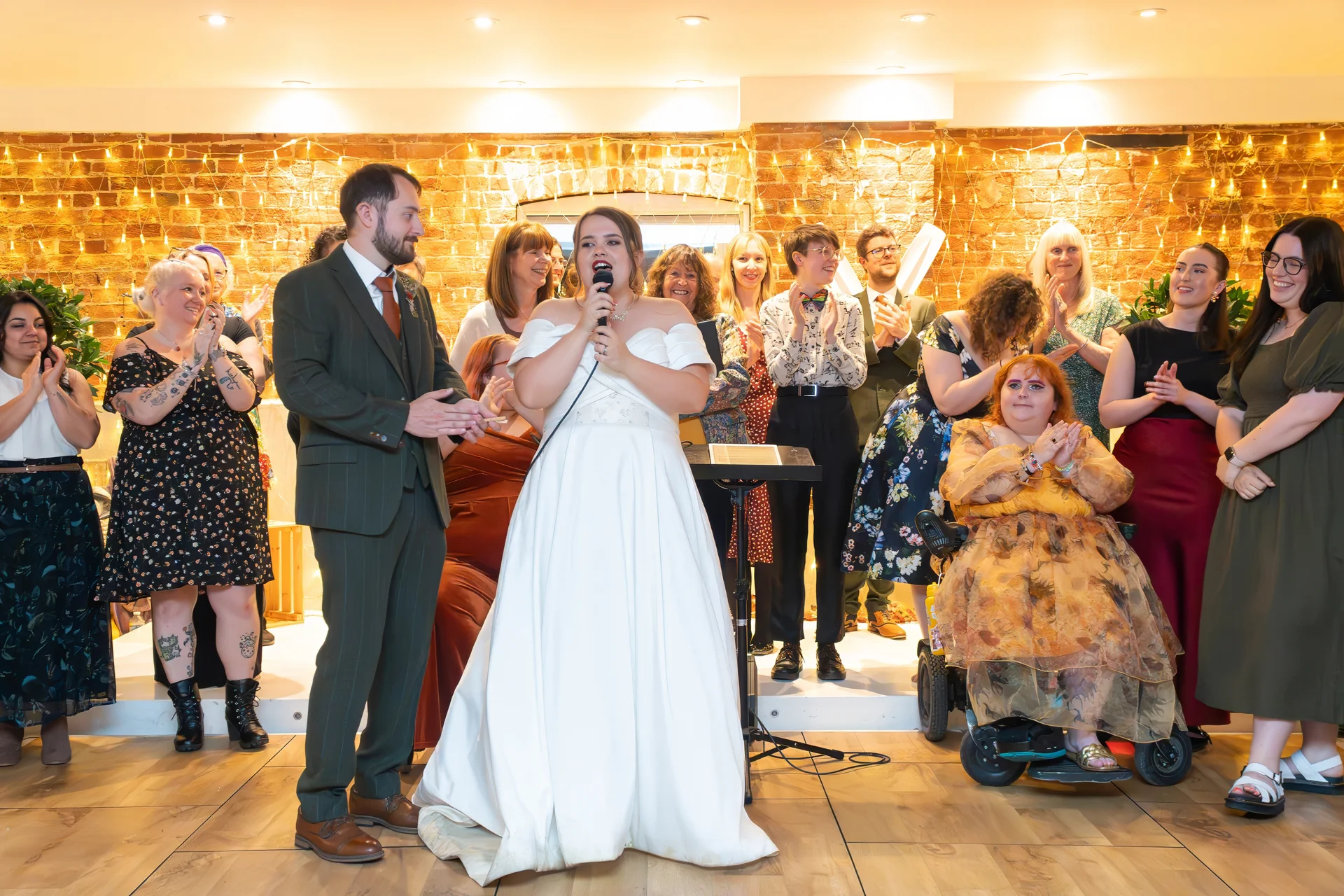 A bride in a white off-the-shoulder gown speaks into a microphone during her wedding reception, flanked by her partner in a green suit and cheering guests against an exposed brick wall lit with fairy lights.