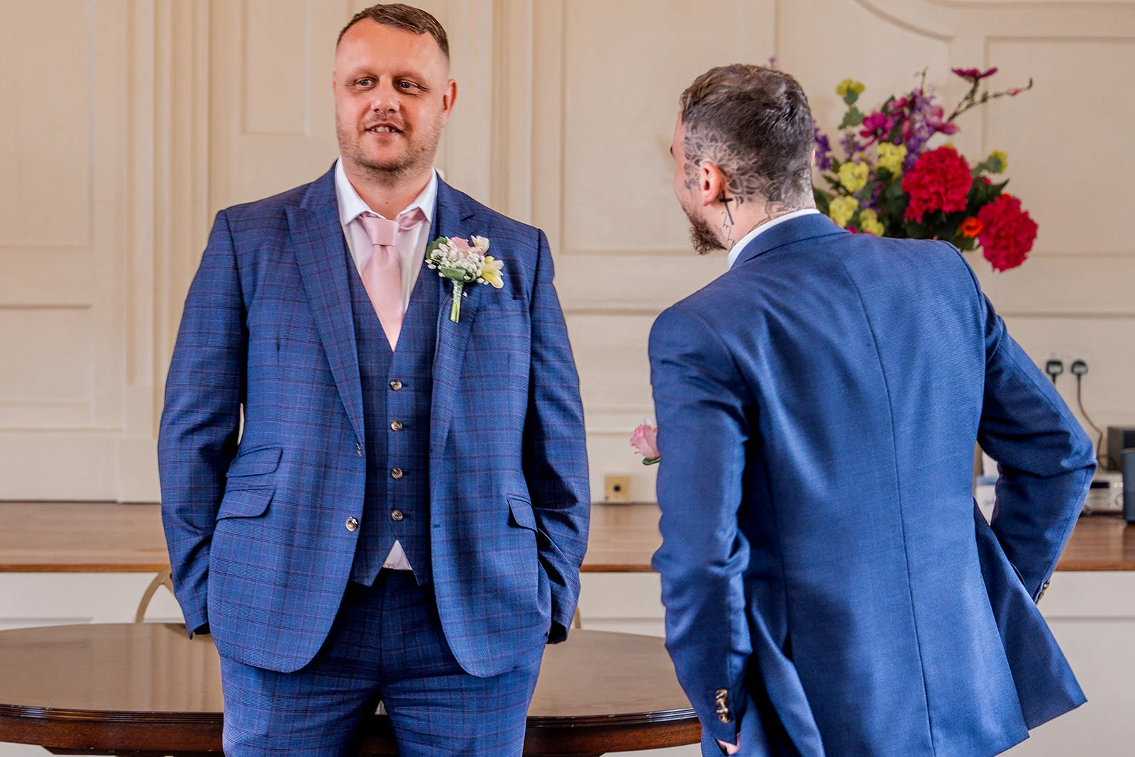 Two grooms in matching blue suits stand smiling indoors next to a table with vibrant floral arrangements.