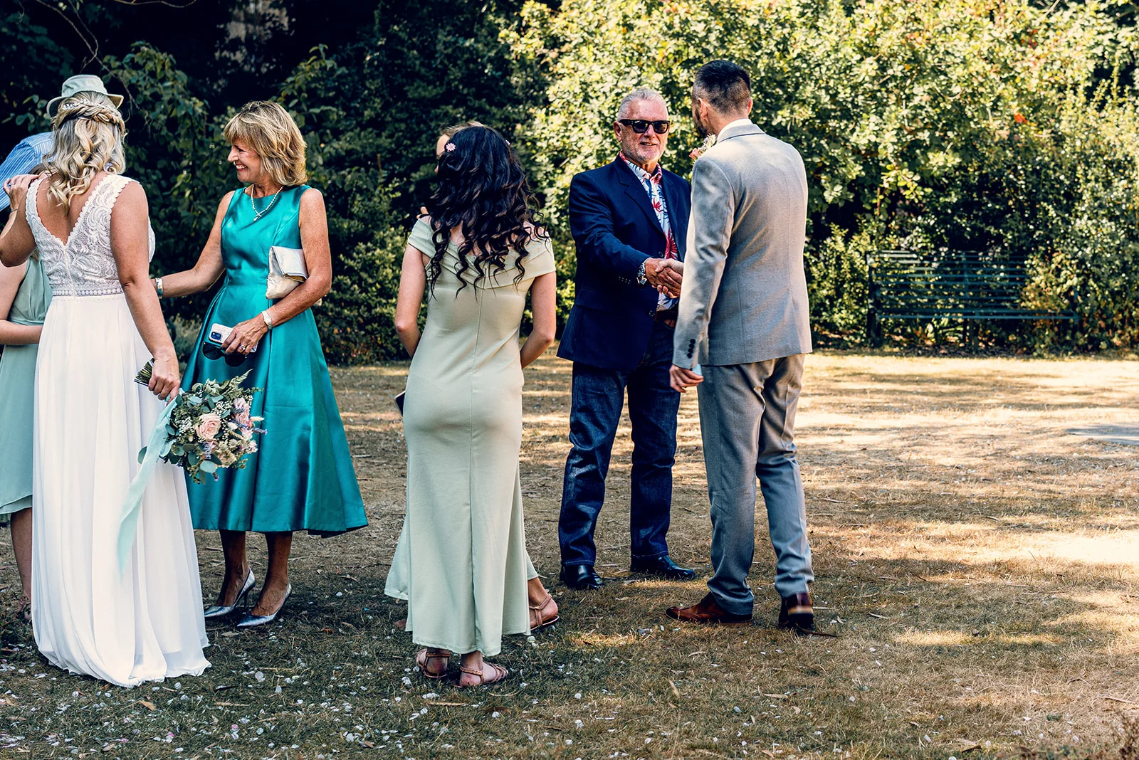 The groom in a grey suit shakes hands with an older guest during an outdoor garden wedding reception while the bride and guests look on.