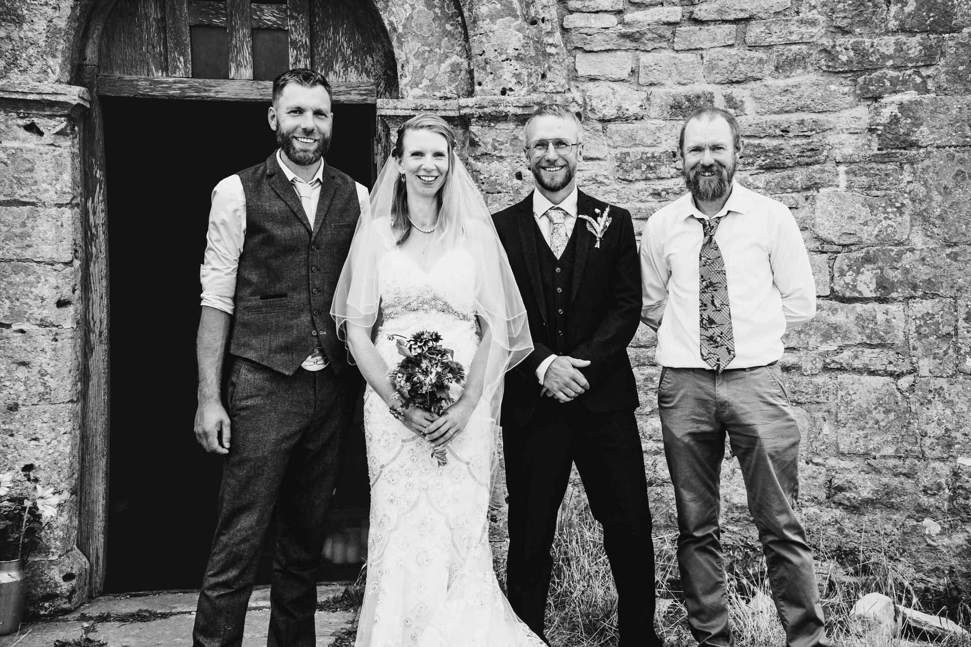 A smiling bride and groom pose outside a historic stone church doorway alongside two groomsmen in this classic black and white portrait.