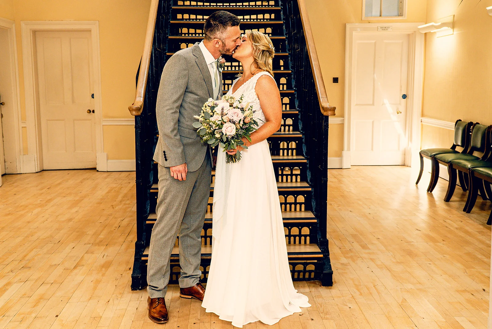 A newly married couple share a tender kiss on a wooden floor at the foot of an ornate staircase inside a venue.